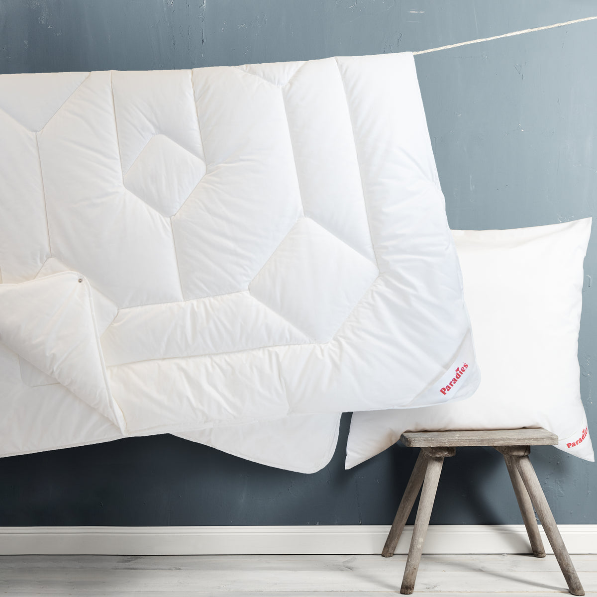 Zwei weiße Steppdecken mit geometrischem Muster, eine hängt, die andere liegt auf einem kleinen Holzschemel vor einer dunkelblauen Wand