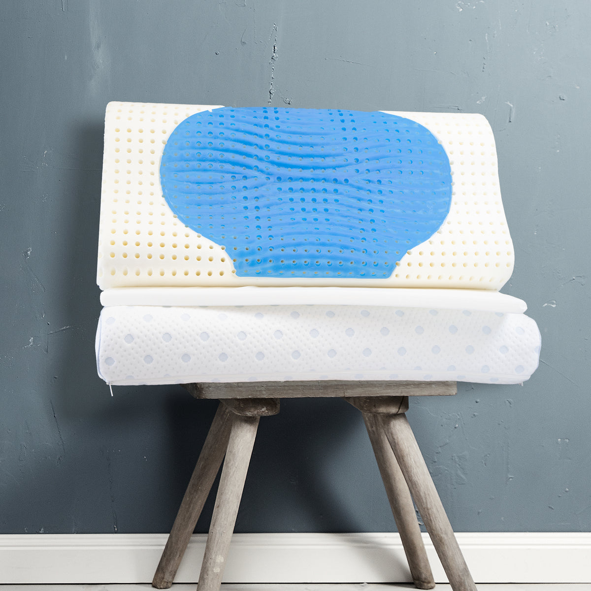 Ein weißes ergonomisches Kissen mit blauer Gelauflage und kleinen Löchern, auf einem hölzernen Hocker vor grauer Wand.
