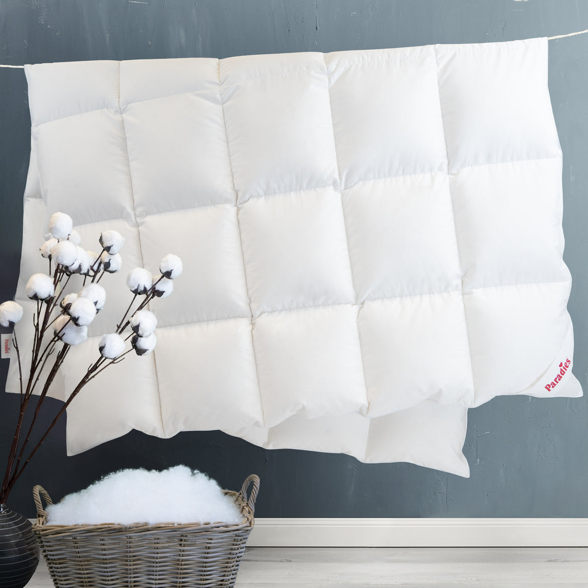 Weiße Steppdecke mit quadratischer Steppung an einer grauen Wand hängend, davor ein Korb mit Füllmaterial und ein Zweig mit Baumwollblüten