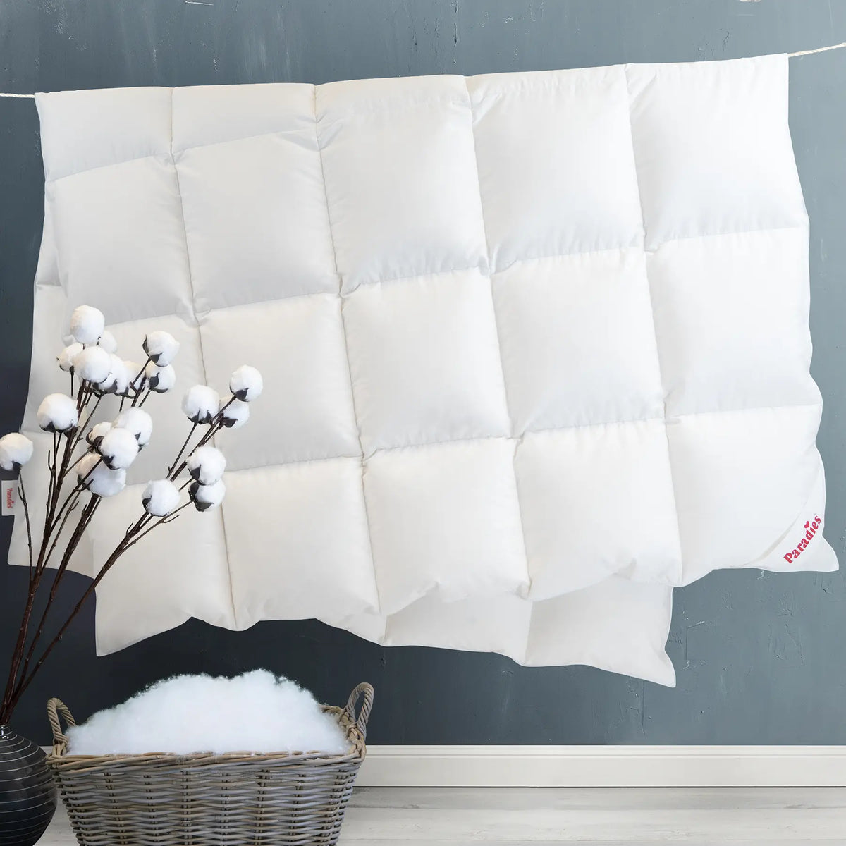 Weiße Steppdecke mit quadratischem Muster und rotem Schriftzug 'Paradies' in der Ecke, vor dunkelgrauer Wand und Korb mit Füllmaterial