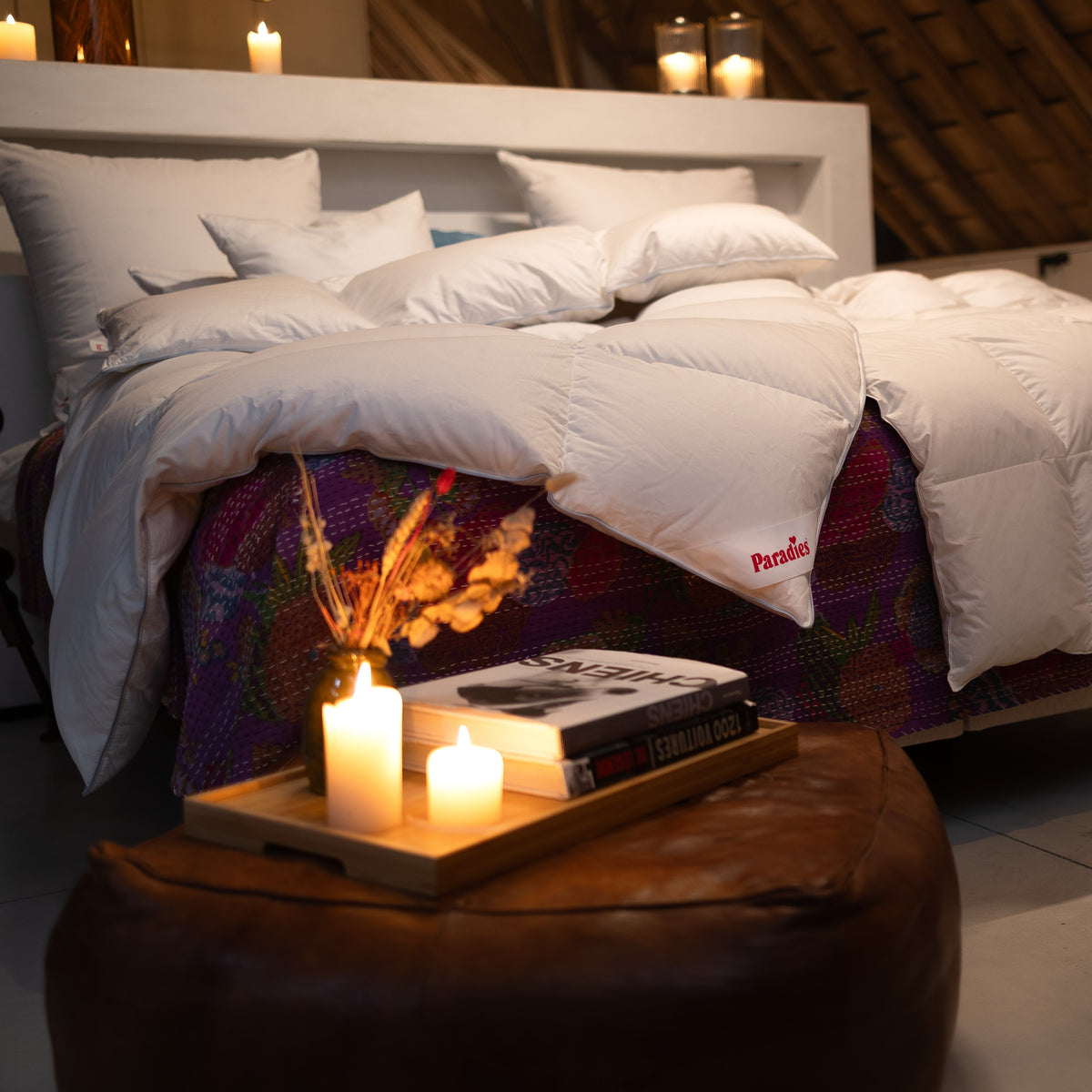 Bett mit weißer Daunendecke und Kissen auf bunter Decke, davor ein brauner Hocker mit Holztablett, darauf getrocknete Blumen, Bücher und brennende Kerzen