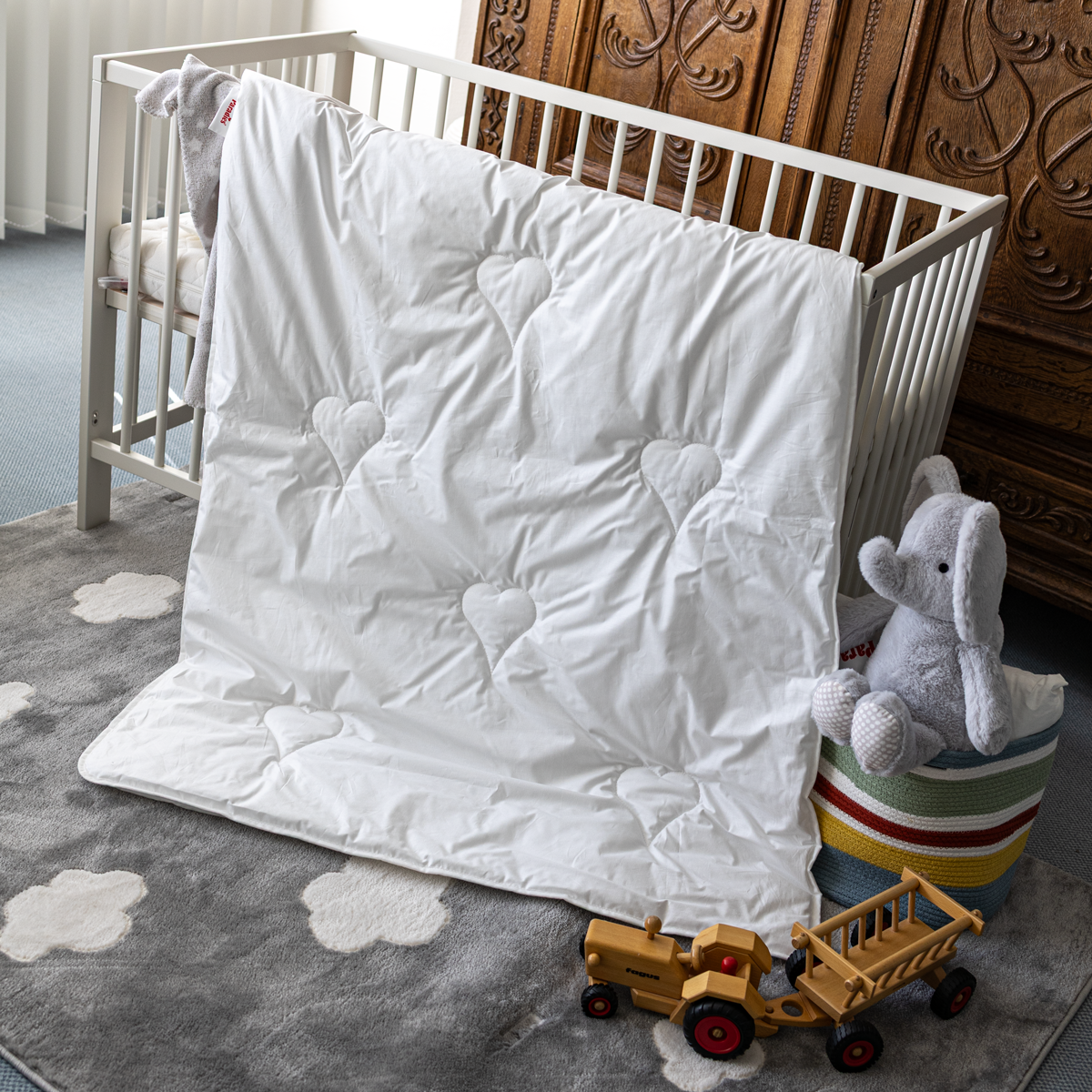 Weiße Ganzjahresbettdecke mit Herzmustern lehnt an einem weißen Kinderbett auf einem grauen Teppich mit Wolkenmotiven, daneben ein grauer Plüschelefant auf einem bunten Sitzhocker und ein Holzspielzeugtraktor