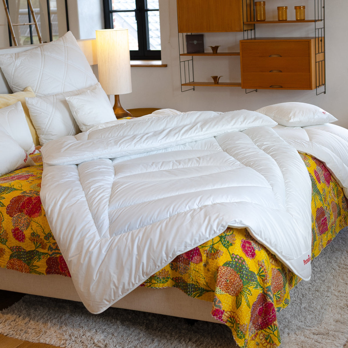 Weiße Avera Ganzjahresdecke liegt auf einem Bett mit gelber, gemusterter Bettdecke in einem hellen Schlafzimmer mit Holzregal und Lampe.