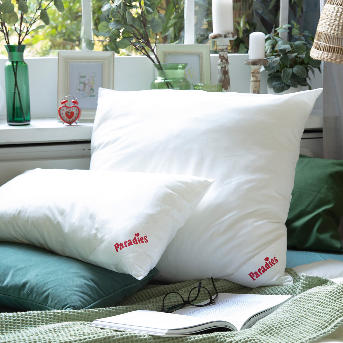 Zwei weiße Kissen mit rotem Schriftzug 'Paradies' liegen auf einem grünen Bett mit offener Zeitschrift und Brille davor