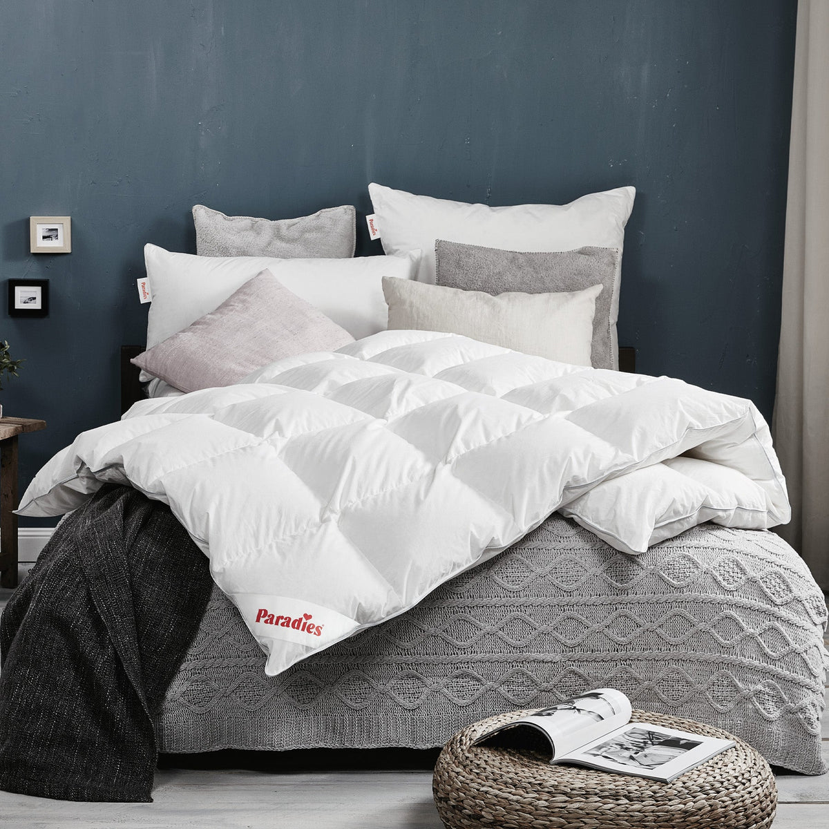 Bett mit weißer Steppdecke, mehreren Kissen in Weiß und Grau sowie grauer Strickdecke vor dunkelgrauer Wand