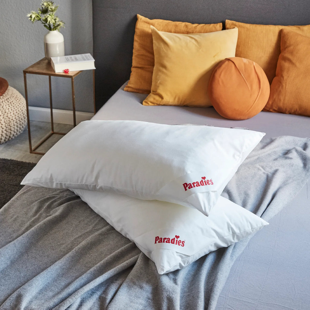Zwei weiße Kissen mit roter Aufschrift 'Paradies' liegen auf einem hellgrauen Bett mit orangefarbenen und gelben Kissen im Hintergrund