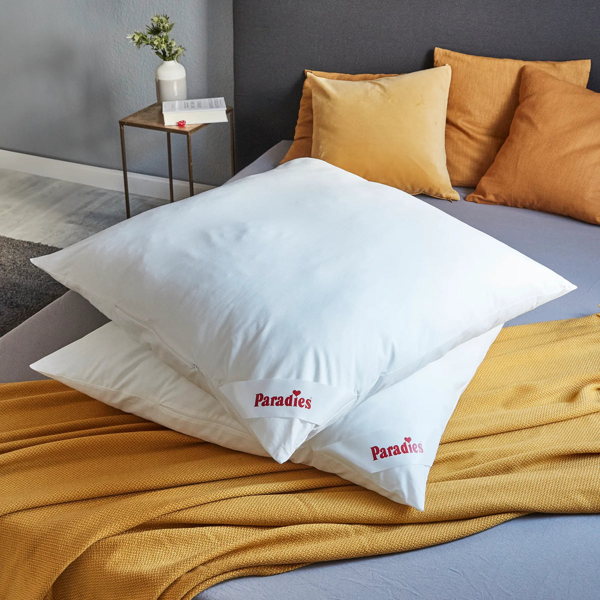 Zwei weiße Kissen mit rotem Paradies-Logo liegen auf einer gelben Decke auf einem Bett mit gelben und orangefarbenen Kissen im Hintergrund