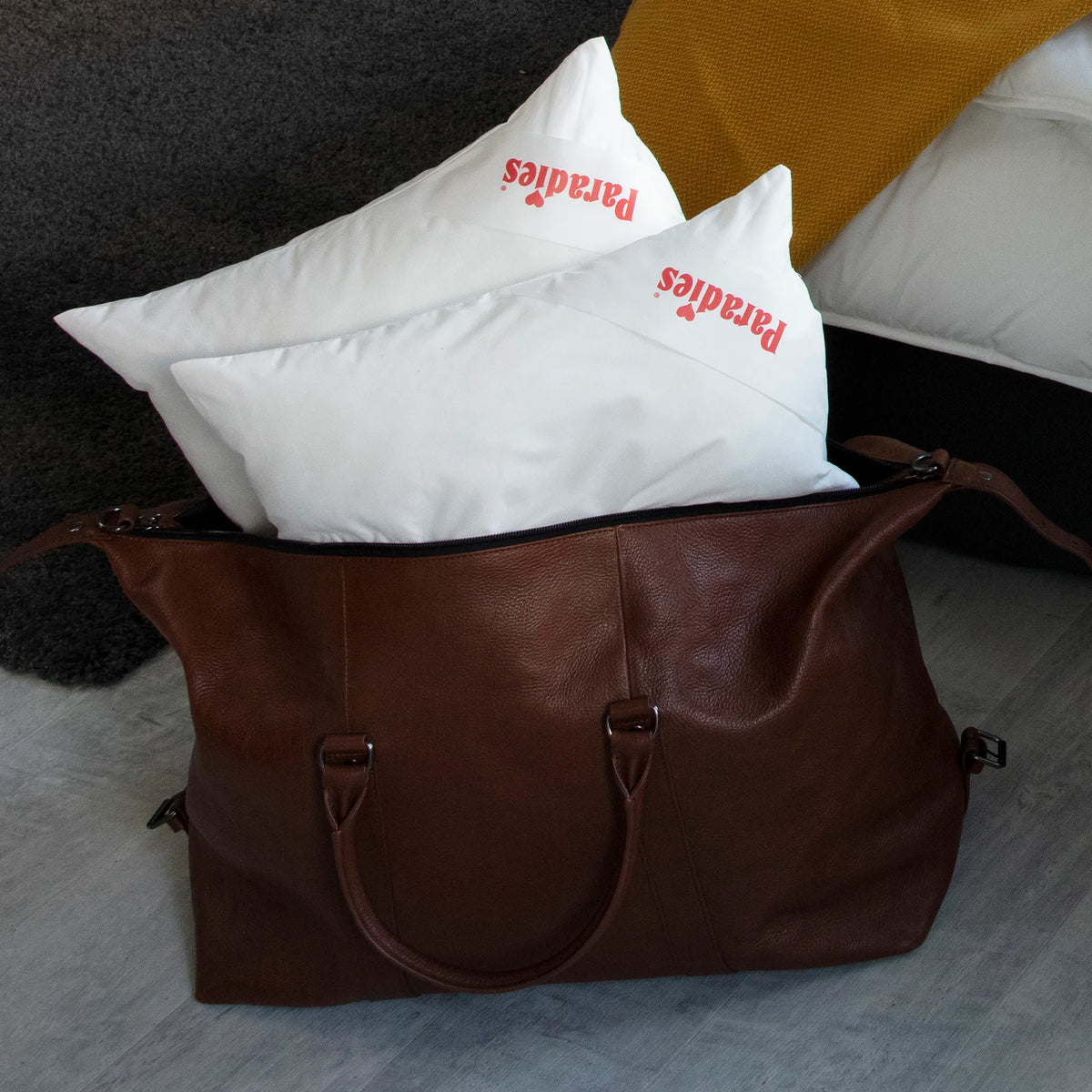Braune Ledertasche mit zwei weißen Kissen mit rotem Schriftzug 'paradies' auf grauem Boden vor gelbem Kissen