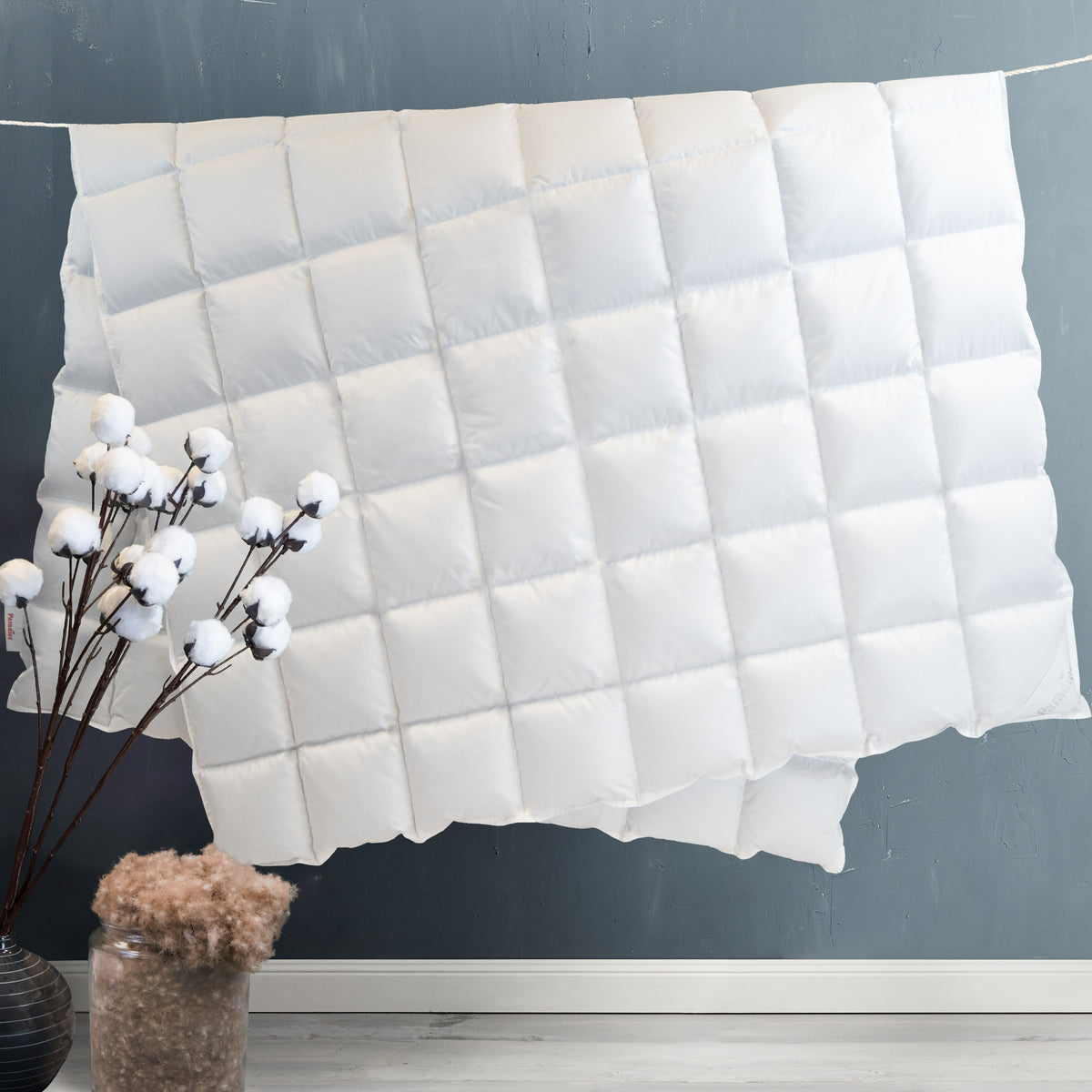 Weiße, quadratisch gesteppt wirkende Decke an einer dunkelgrauen Wand mit zwei Vasen davor, eine mit Baumwollzweigen, die andere mit braunem Deko-Material