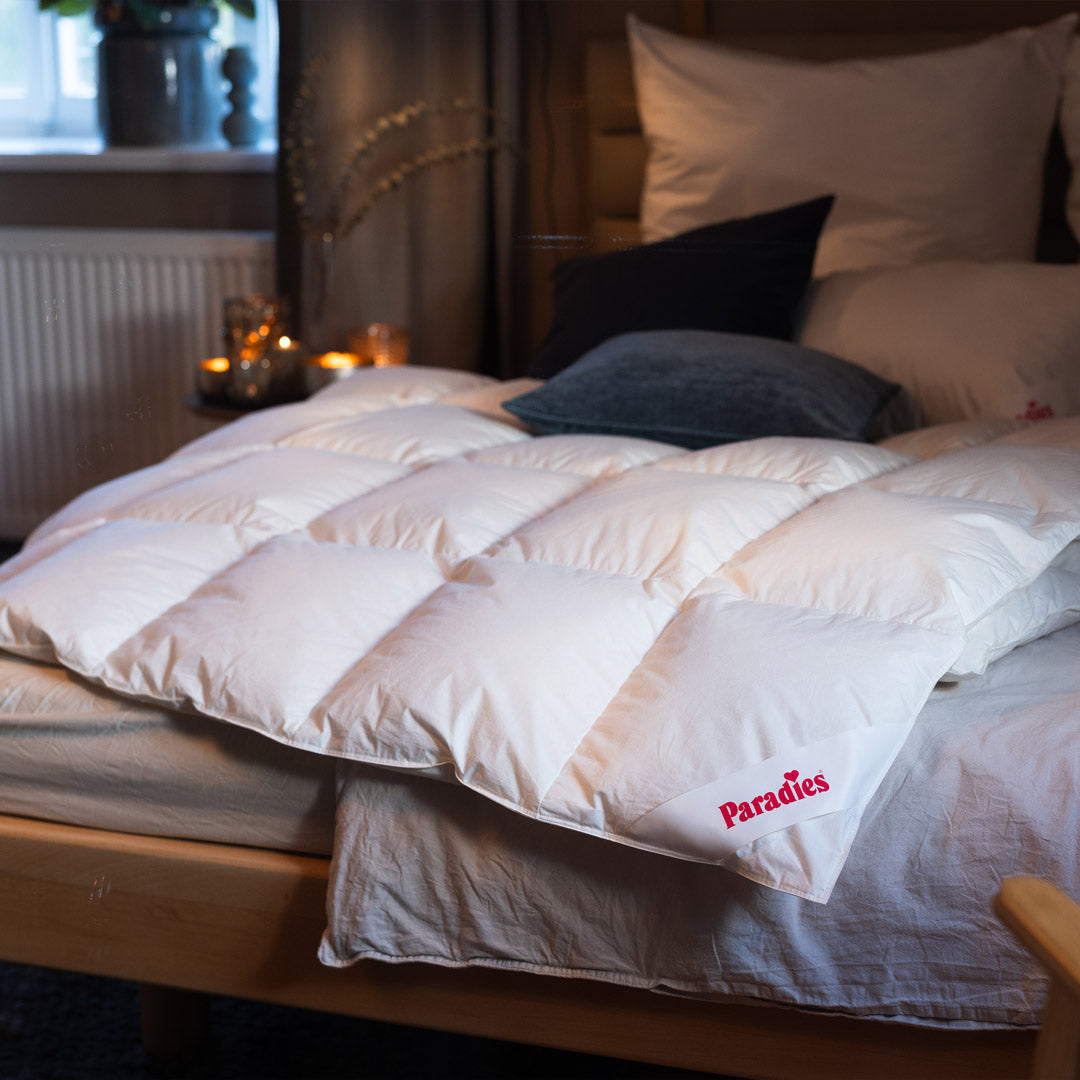 Bett mit weißer Steppdecke, auf der Ecke ein rotes Logo mit dem Schriftzug 'Paradies', im Hintergrund Kissen und gedämpfte Beleuchtung