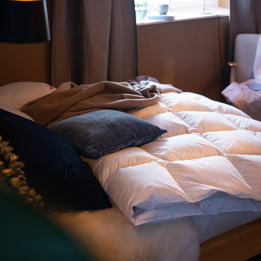 Unordentlich gemachtes Bett mit Daunendecke, mehreren Kissen und einer Decke in einem gemütlichen Zimmer mit Fenster im Hintergrund