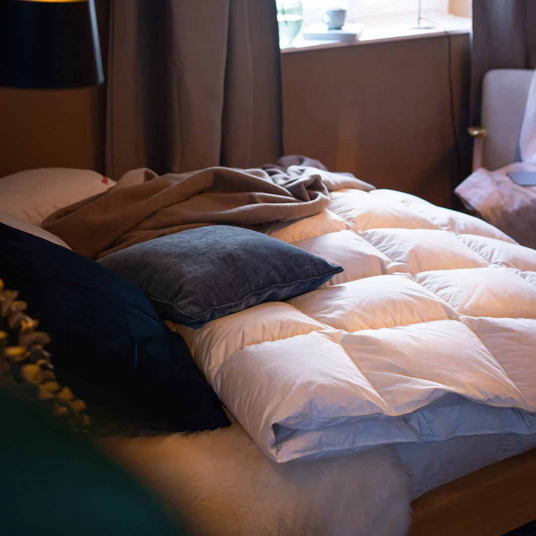 Unordentlich gemachtes Bett mit mehreren Kissen und einer Decke vor einem Fenster mit Vorhängen