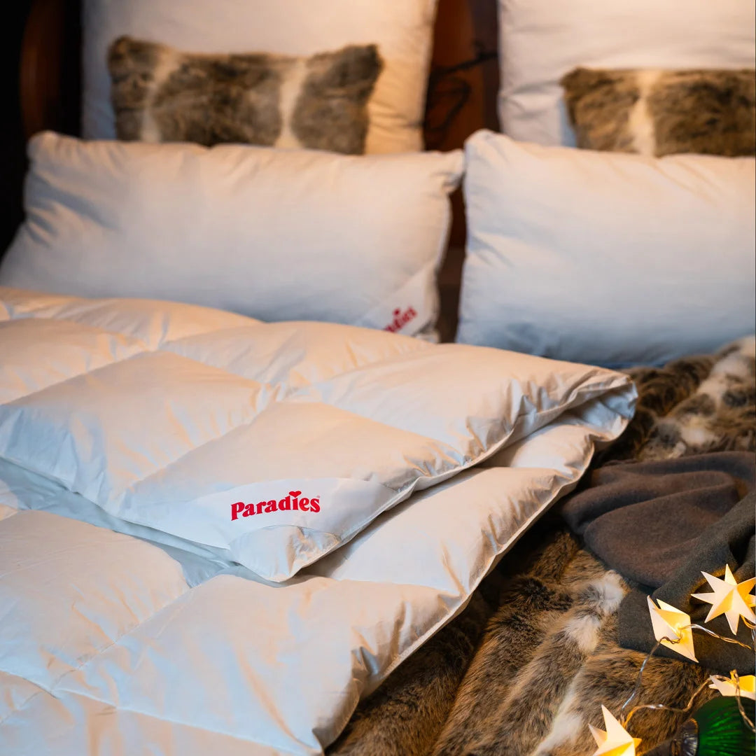 Zwei weiße Kissen und eine weiße Daunendecke mit rotem Paradies-Logo liegen auf einem Bett mit braunem Fellüberwurf und zwei Kissen mit Fellbezug
