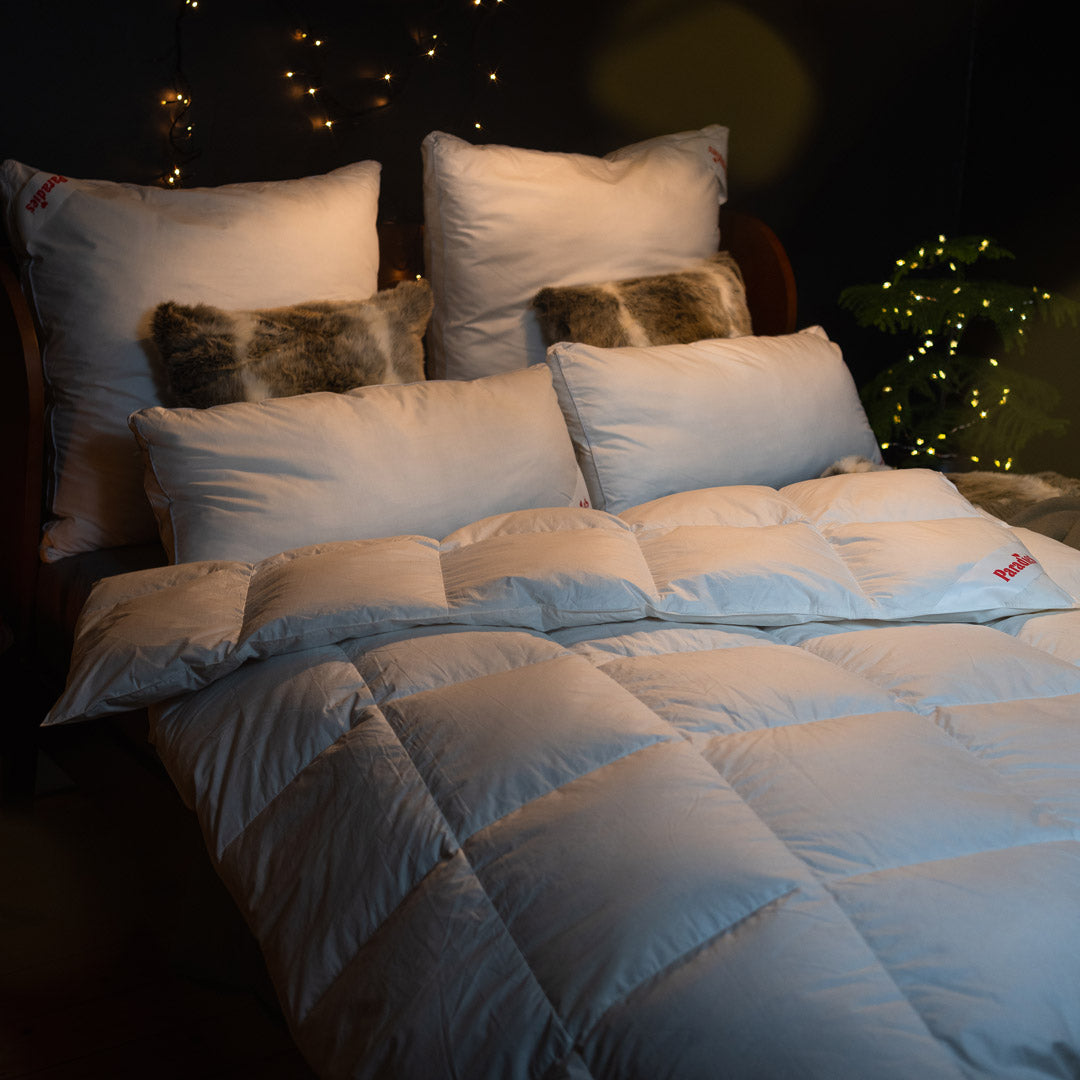 Bett mit mehreren weißen Kissen und einer weißen Steppdecke in einem dunklen Raum mit Lichterkette im Hintergrund