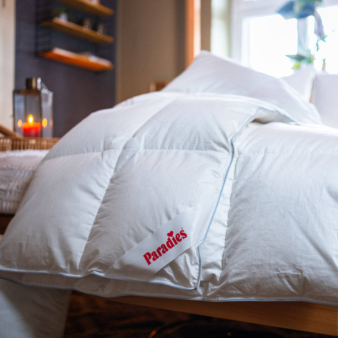 Nahaufnahme einer weißen Bettdecke mit einem eckigen Label mit roter Aufschrift 'Paradies' auf einem Bett in einem gemütlichen Schlafzimmer