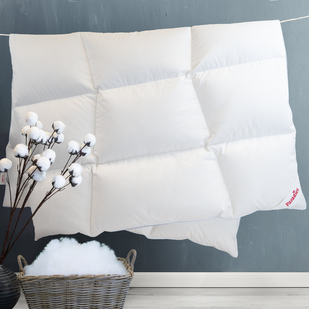 Zwei weiße, gesteppte Kissen mit rotem Paradies-Logo liegen vor einer grauen Wand, daneben ein Korb mit weißer Füllung und ein Zweig mit Baumwollblüten.