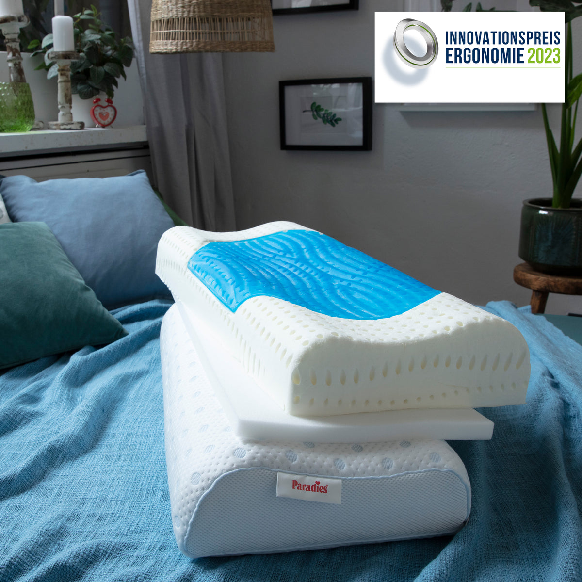 Kissen liegen übereinander auf einem blauen Bettbezug in einem Schlafzimmer mit Kerze, Pflanzen und Wandbildern