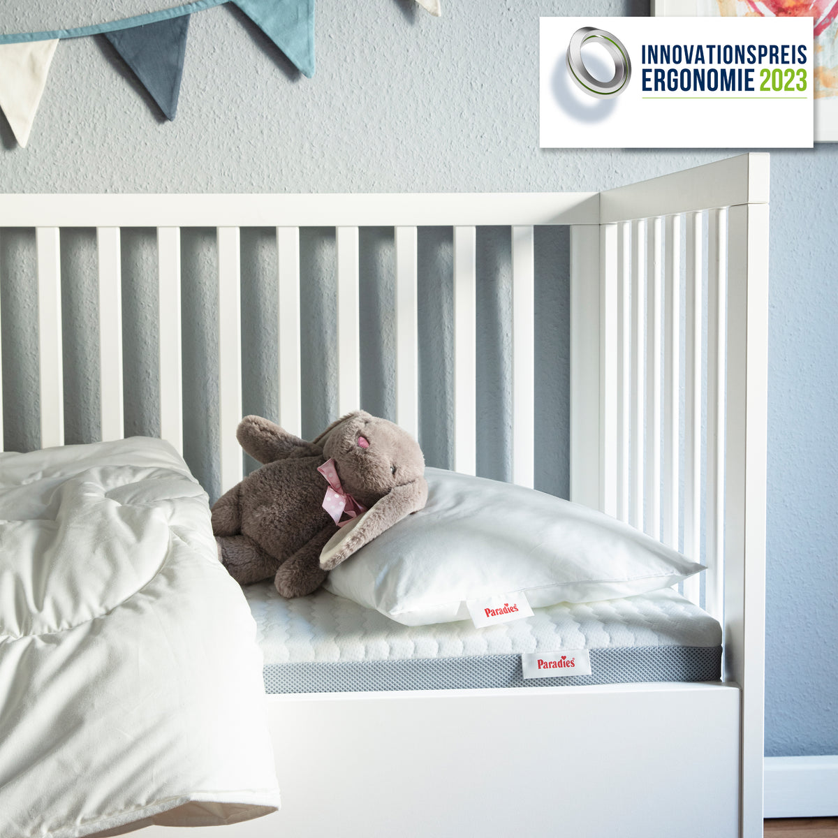 Weißes Babybett mit Kuscheltier auf Kissen und Decke vor hellblauer Wand mit Wimpelkette