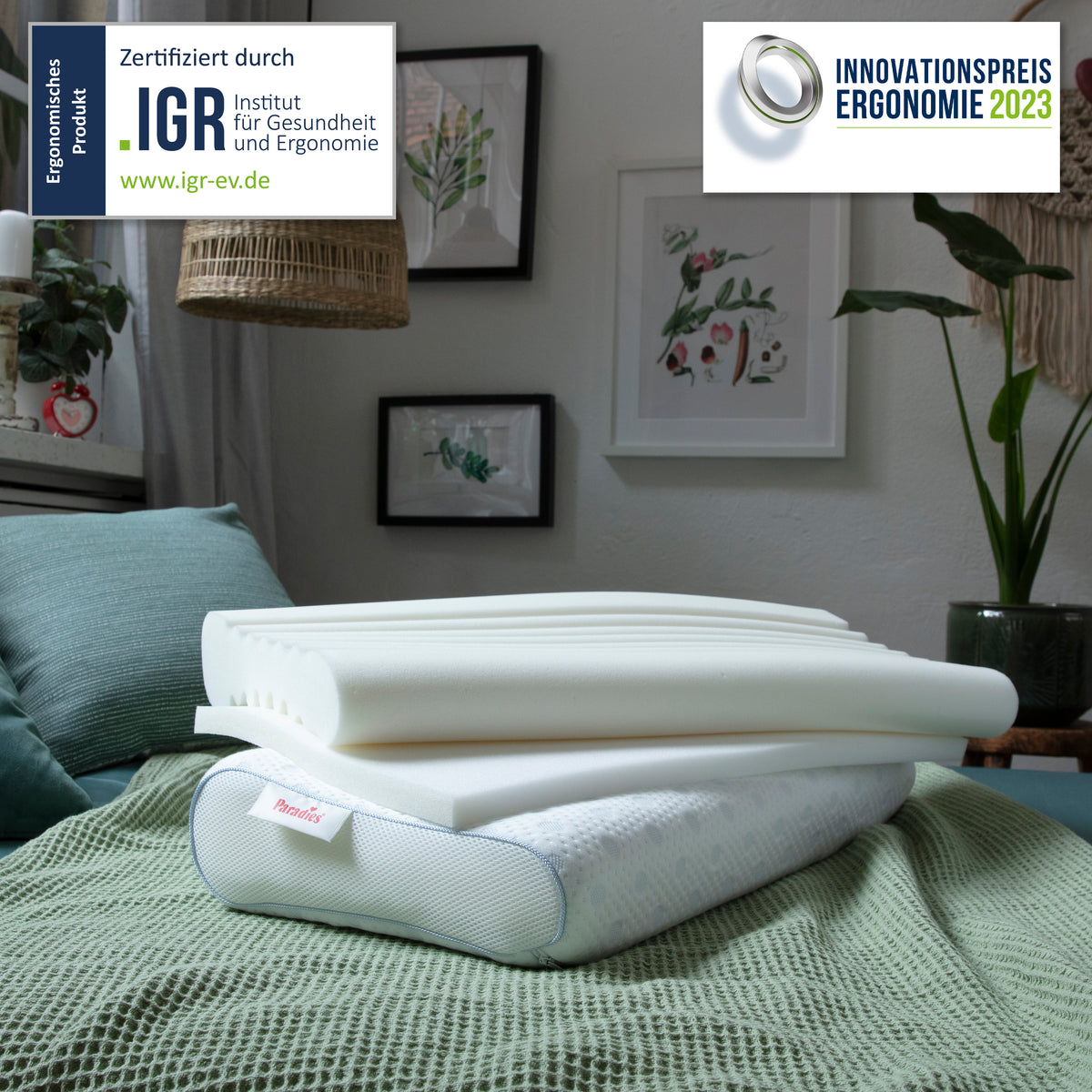 Auf einem grünen Bett liegt ein ergonomisches Kissen mit aufgeschichteten weißen Schaumstoffeinlagen, im Hintergrund sind eingerahmte Pflanzenbilder und eine Pflanze zu sehen