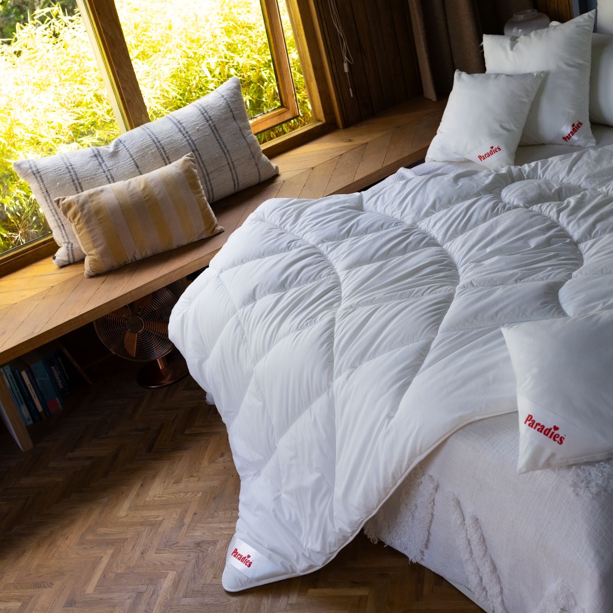 Bett mit weißer Steppdecke und mehreren Kissen mit rotem Schriftzug auf Holzboden neben Fensterbank mit Kissen