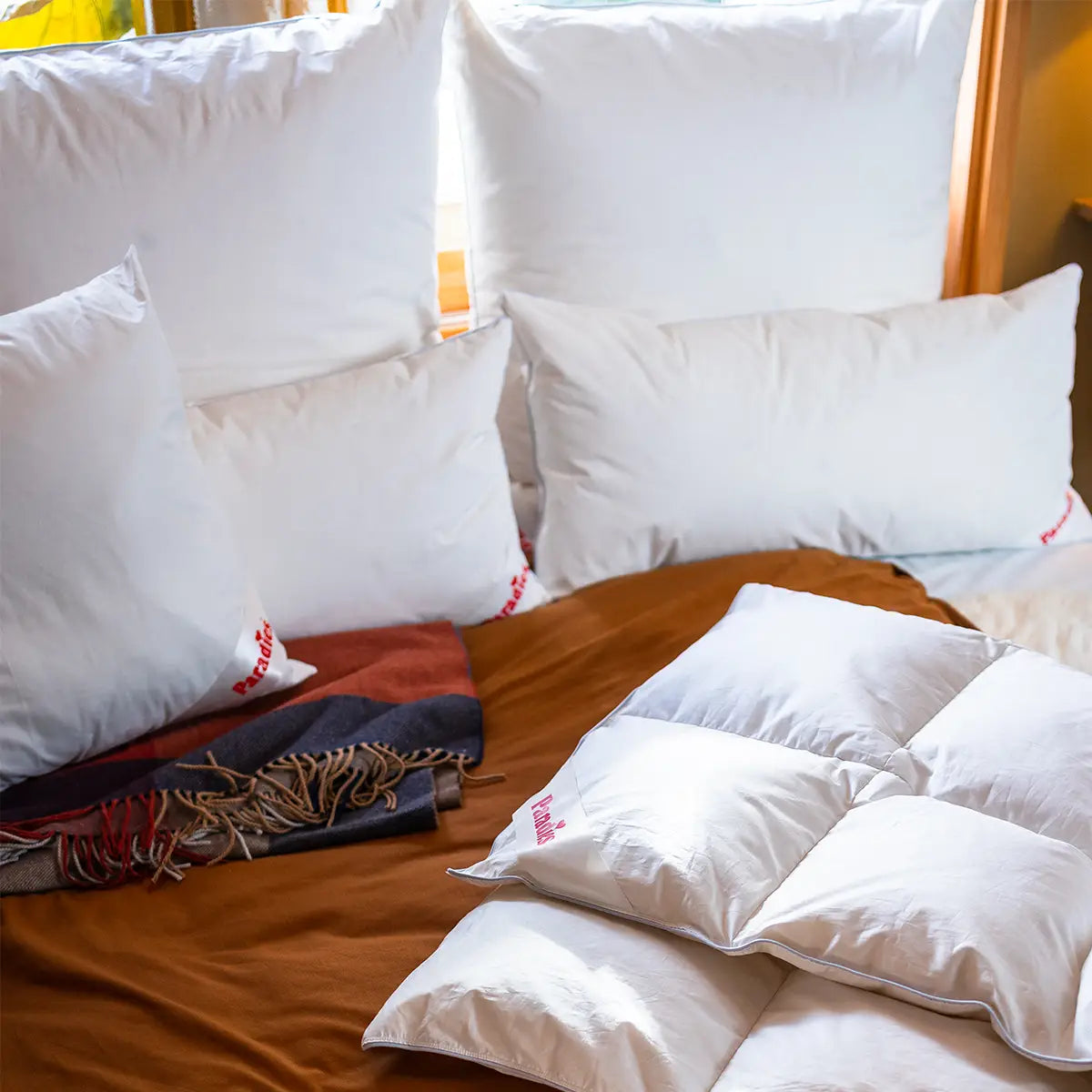 Mehrere weiße Kissen und eine weiße Daunendecke liegen auf einem braunen Bett, daneben liegt ein mehrfarbiger Schal