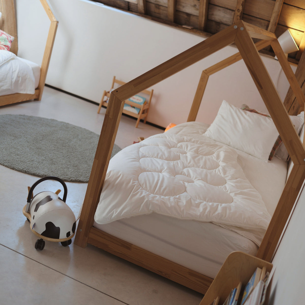 Kinderbett mit Holzrahmen in Hausform, weißer Bettdecke und Kissen neben rundem Teppich und Kinderspielzeug
