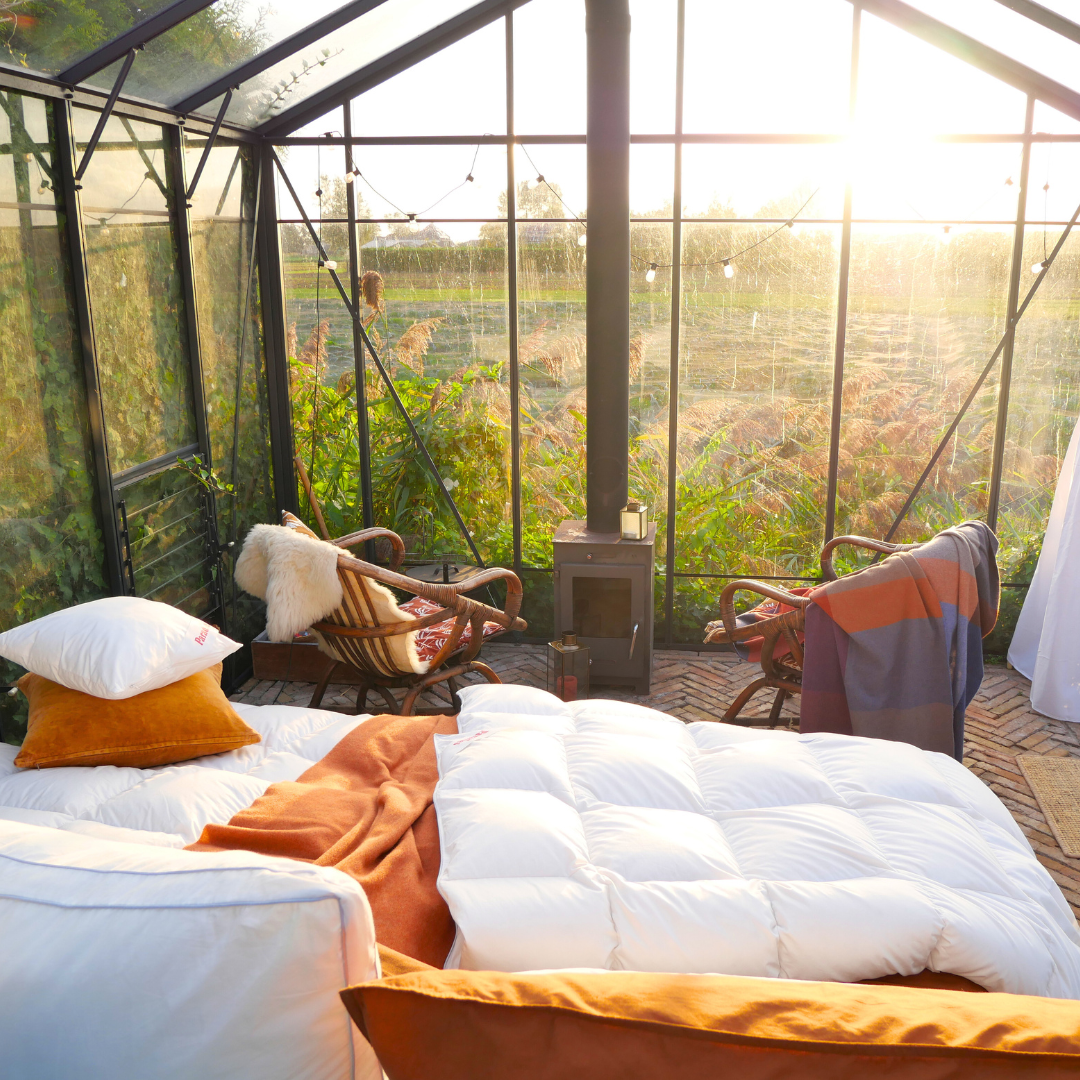 Paradies Winterbettdecke Elma 90 auf Bett in sonnigem Gewächshaus mit zwei Korbsesseln, Kaminofen und orangefarbenen Kissen
