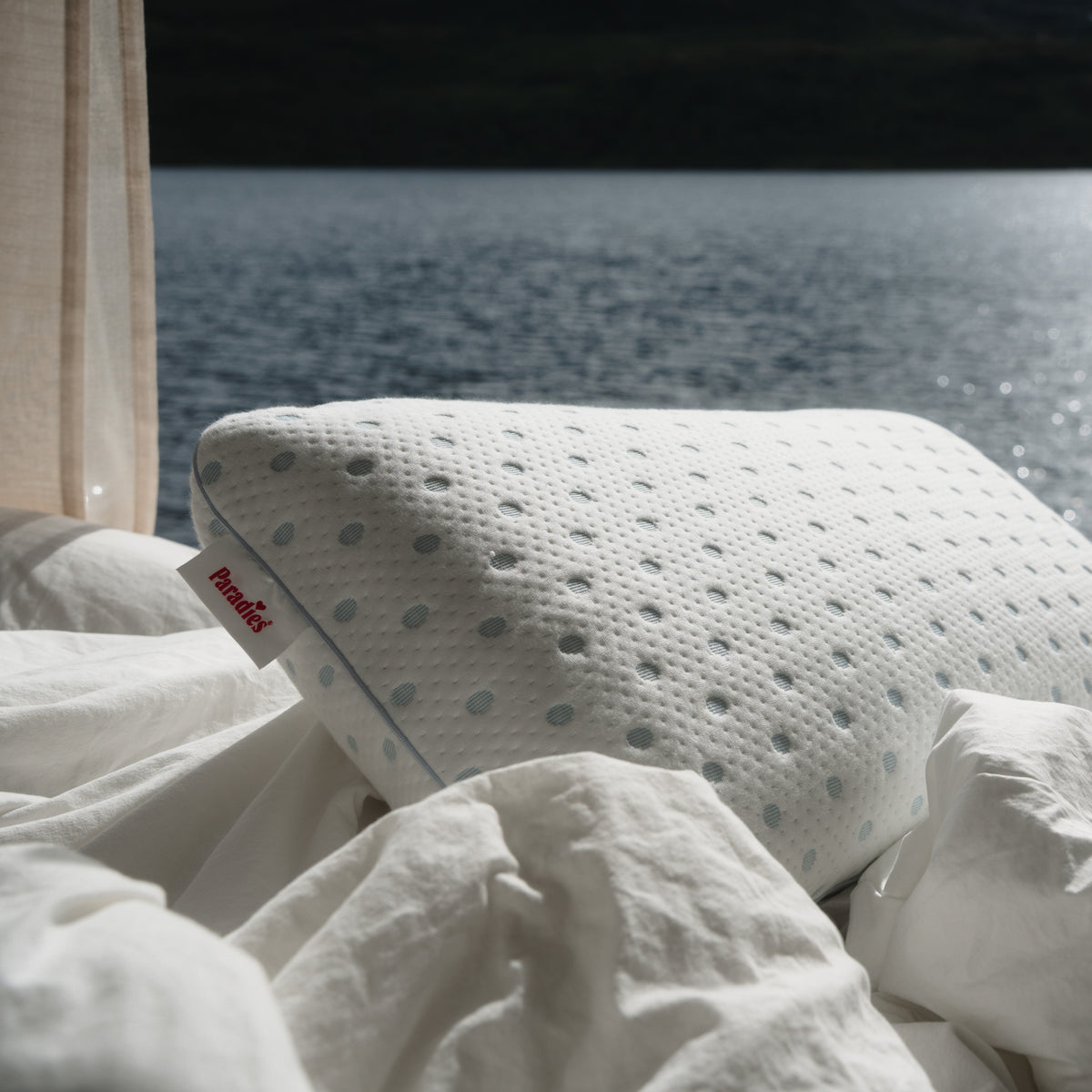 Das weiße Kissen Flexogel Dream mit blauen Punkten liegt auf einem Bett mit weißen Laken. Am Kissenrand steht Paradies. Im Hintergrund ist ein See zu sehen.