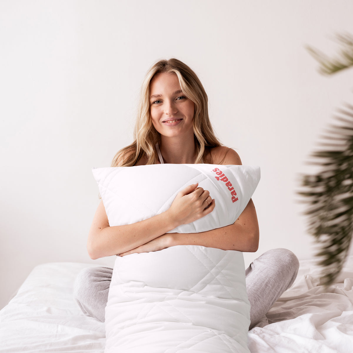 rau sitzt auf einem Bett und hält ein großes weißes Seitenschläferkissen mit rotem Paradies Schriftzug