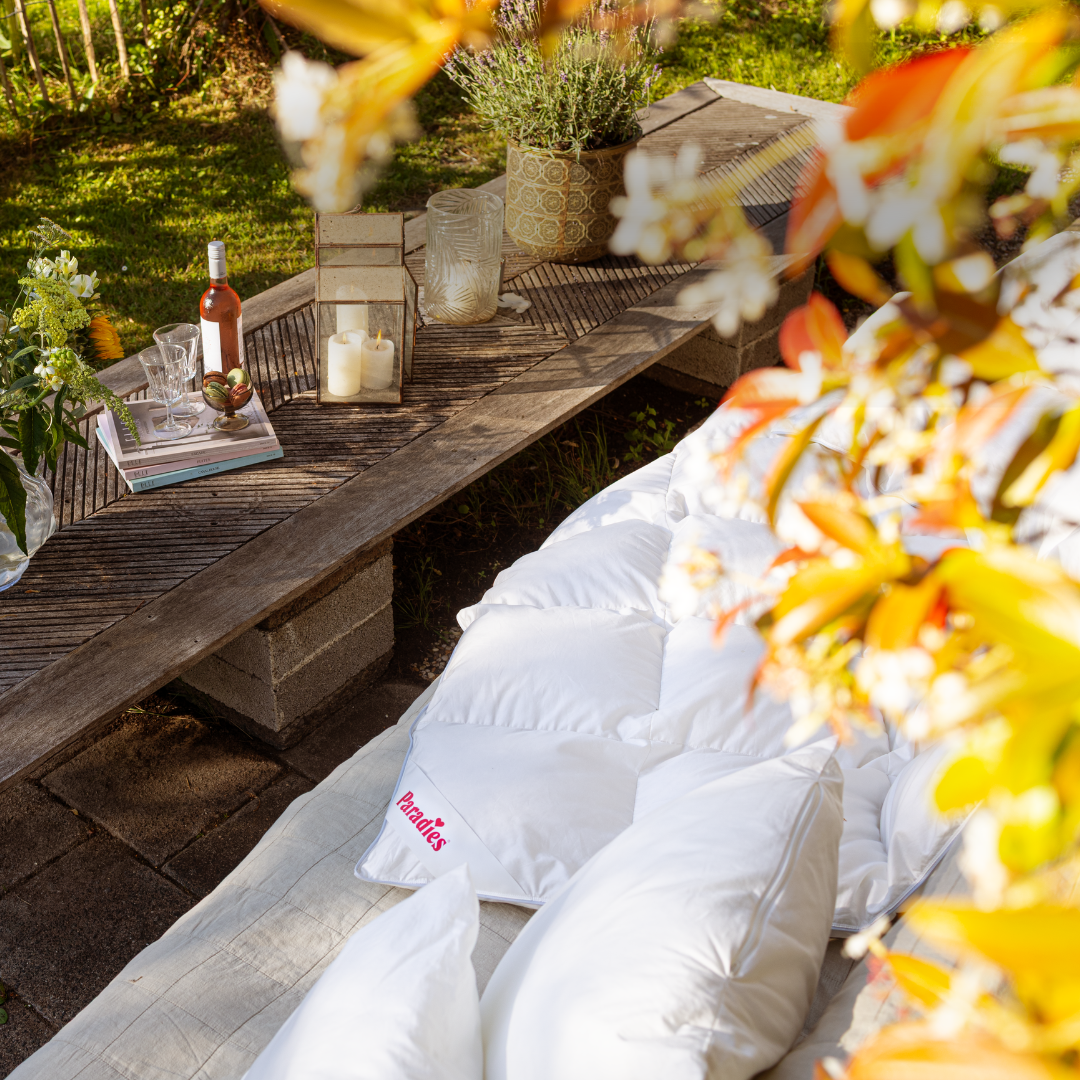 Holztisch im Garten mit Kerzen, Glas, Flasche Roséwein und Büchern, daneben weiße Kissen und Decke mit rotem Schriftzug 'Paradies'