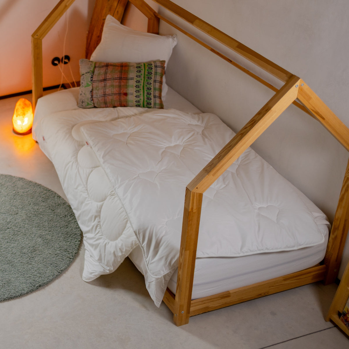 Kinderbett mit Holzrahmen in Hausform, weißer Bettdecke und zwei Kissen neben rundem Teppich und Salzlampe