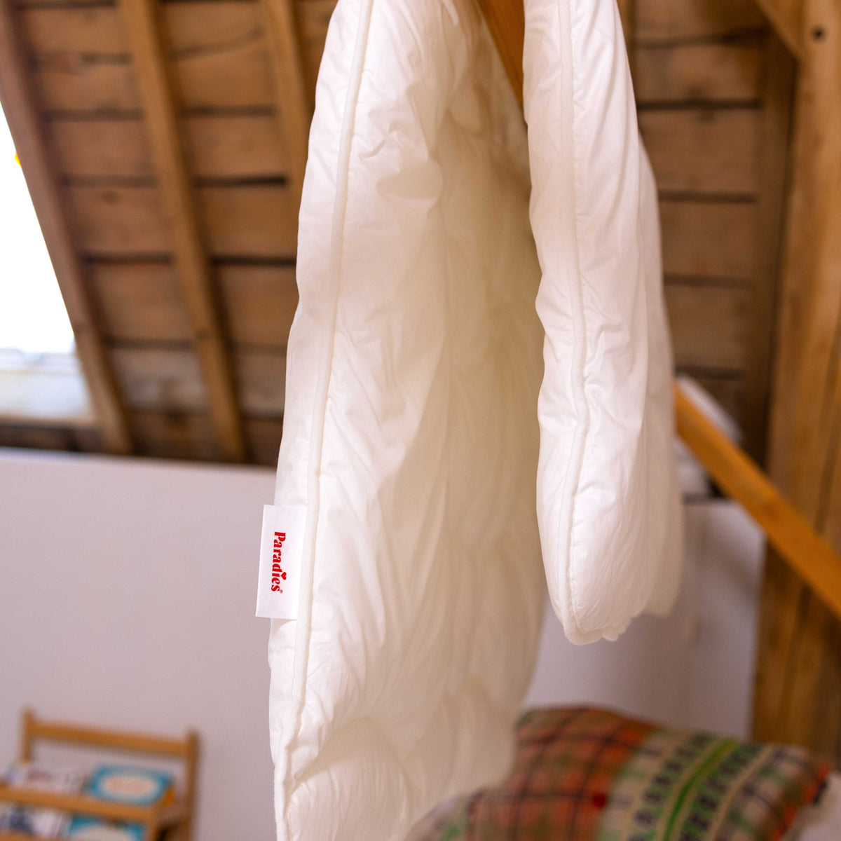 Weiße Kinderdecke Lova mit rotem Etikett 'Paradies' hängt über einem Holzbalken in einem Raum mit sichtbarer Holzbalkendecke