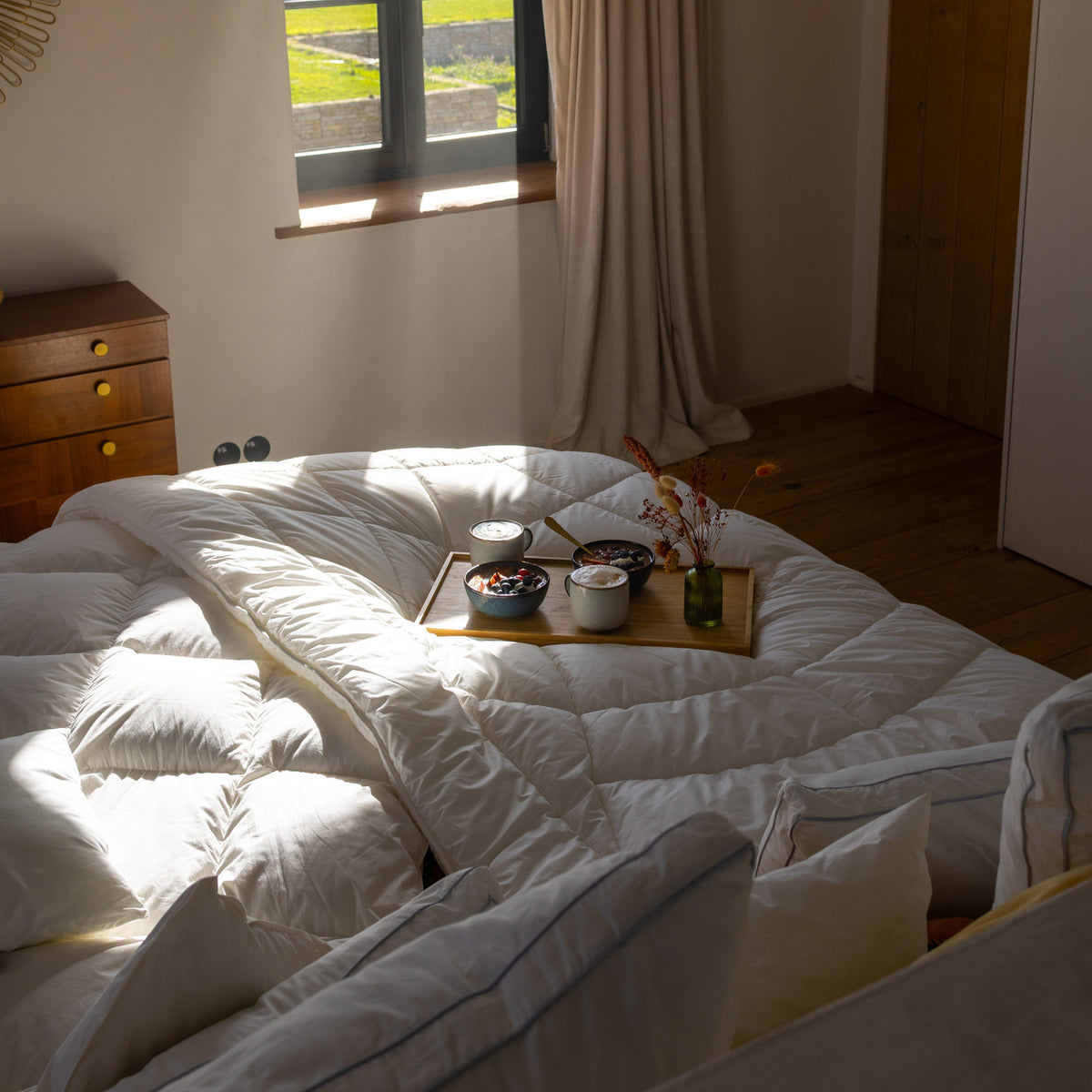 Gemachtes Bett mit weißer Steppdecke und Tablett mit Frühstück vor Fenster mit Vorhang und Holztür im Hintergrund