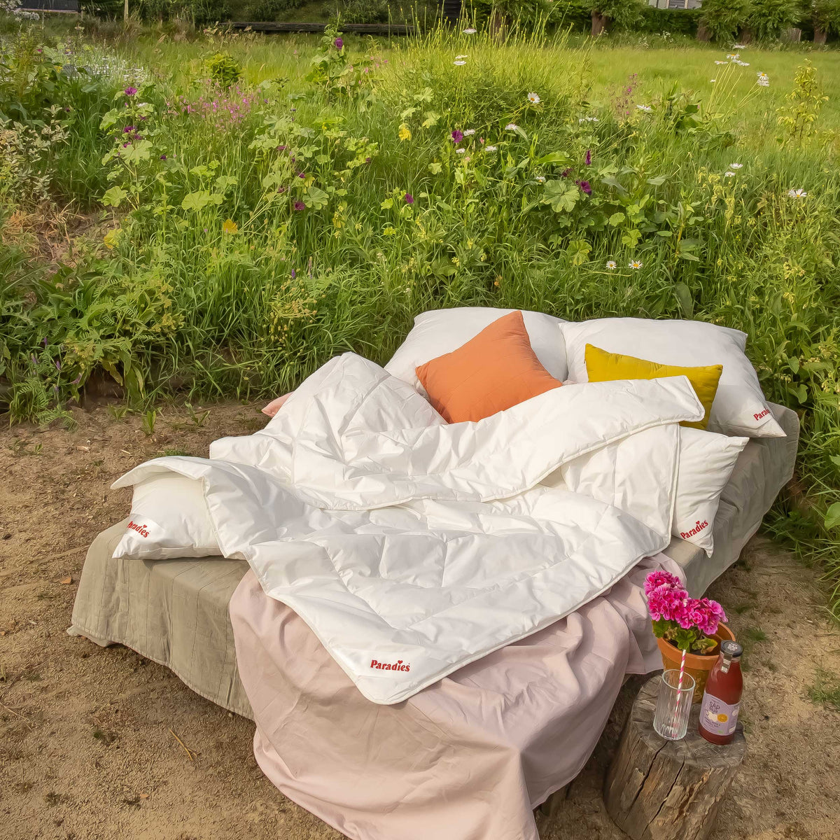 Outdoorbett mit weißer Prima Sommerdecke, orangefarbenem und gelbem Kissen vor einer Wiese mit Wildblumen