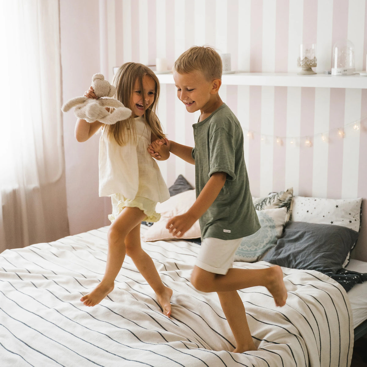 Zwei Kinder springen auf einem gestreiften Bett, eines hält ein Kuscheltier und sie halten sich an den Händen