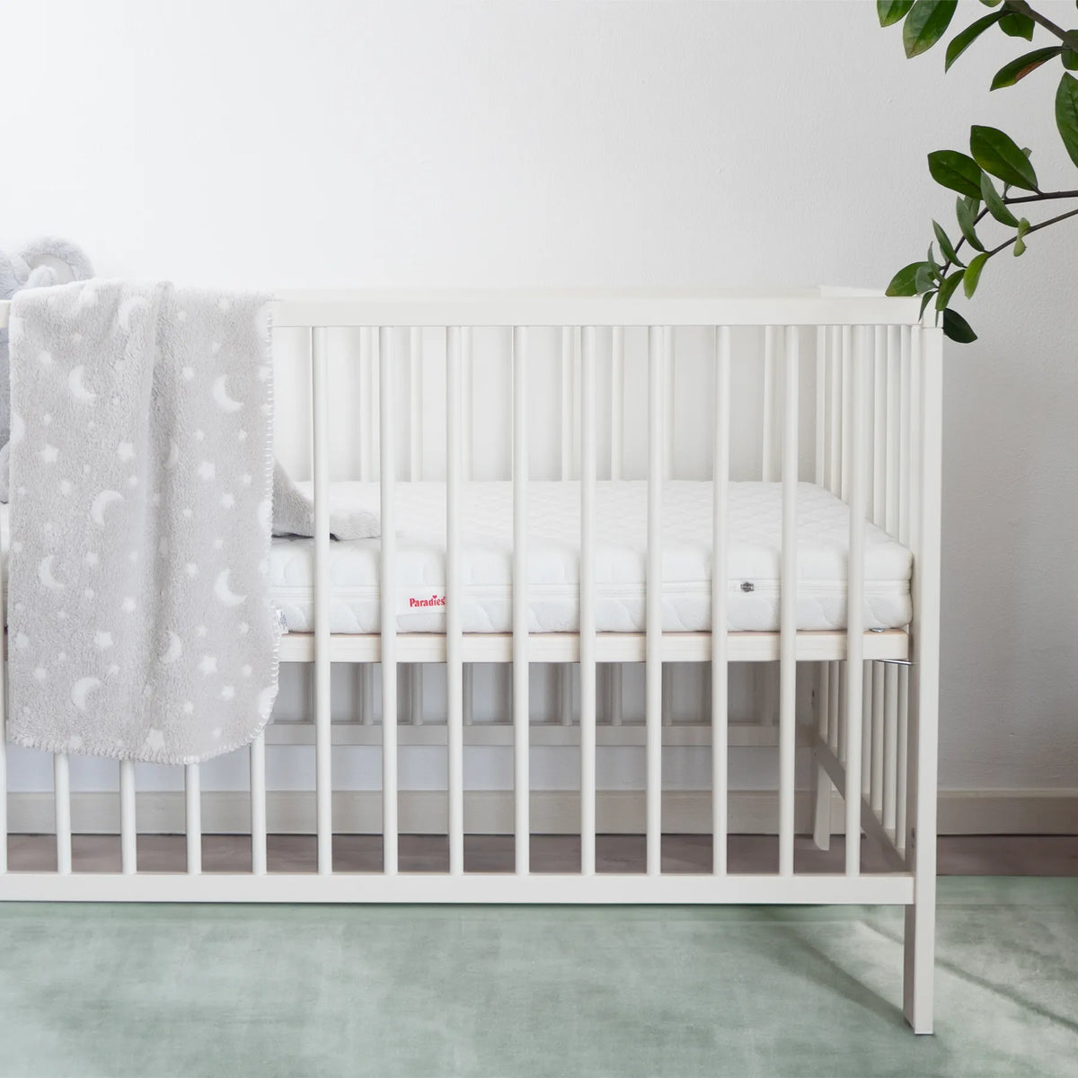 Weißes Babybett mit Gitterseiten, einer weißen Matratze und einer hellgrauen Decke mit Mond- und Sternmuster an der linken Seite