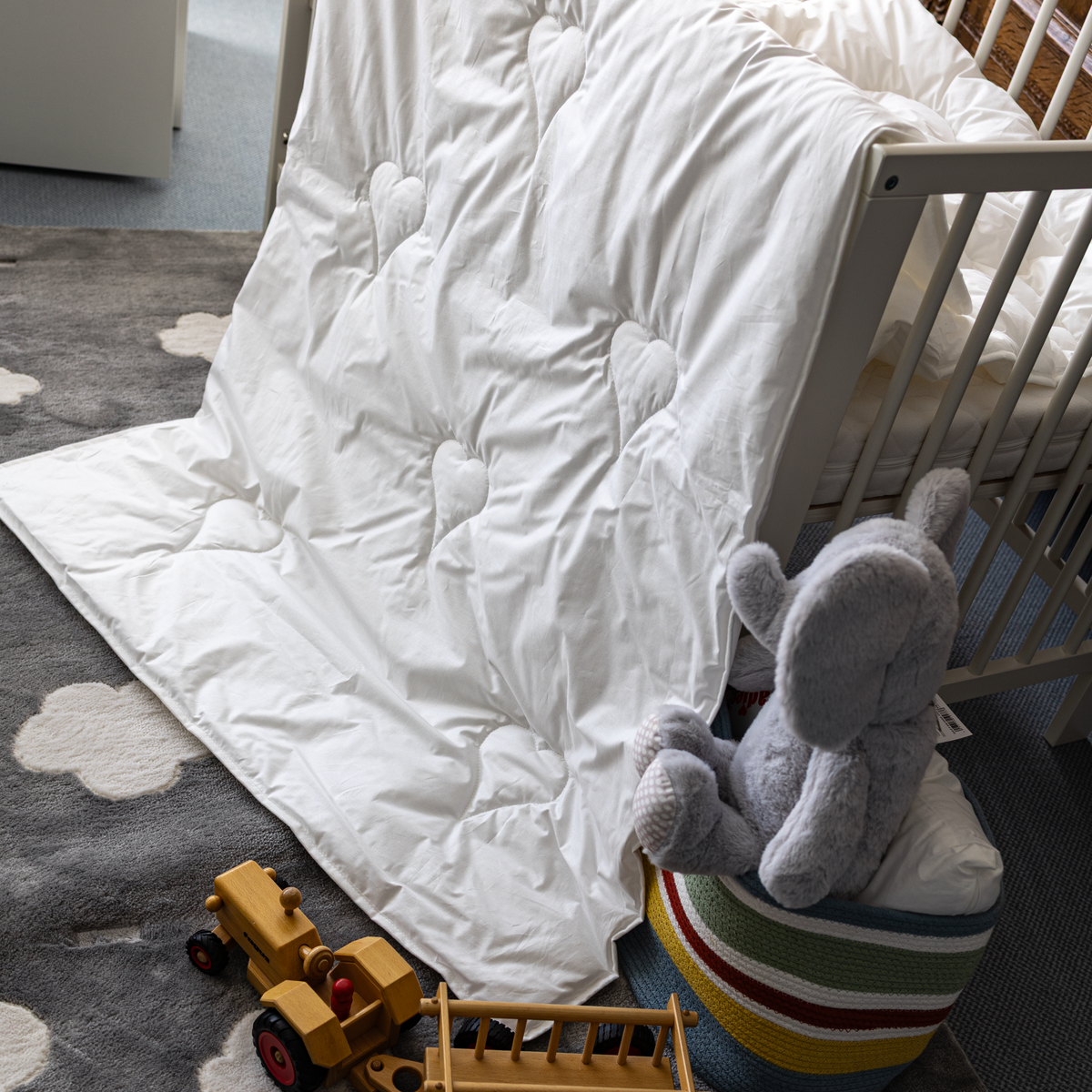 Weiße Ganzjahresbettdecke mit Herzmustern lehnt an einem weißen Kinderbett auf einem grauen Teppich mit Wolkenmotiven, daneben ein grauer Plüschelefant auf einem bunten Sitzhocker und ein Holzspielzeugtraktor