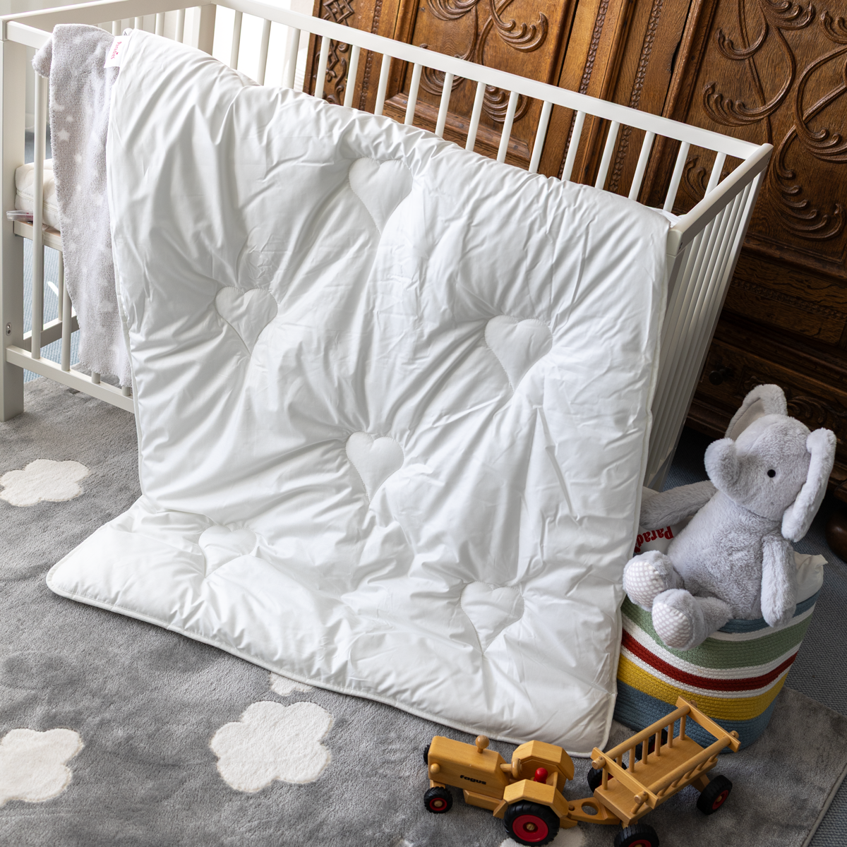 Weiße Ganzjahresbettdecke mit Herzmustern lehnt an einem weißen Kinderbett auf einem grauen Teppich mit Wolkenmotiven, daneben ein grauer Plüschelefant auf einem bunten Sitzhocker und ein Holzspielzeugtraktor