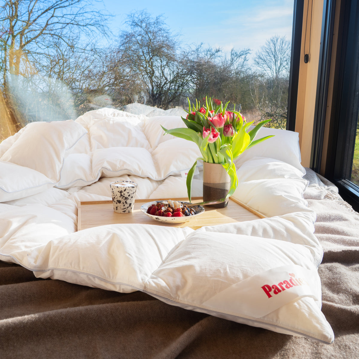 Gemütliches Bett mit weißer Daunendecke, Holztablett mit Tasse, Teller mit Beeren und Vase mit roten Tulpen vor großem Fenster mit Blick auf Bäume