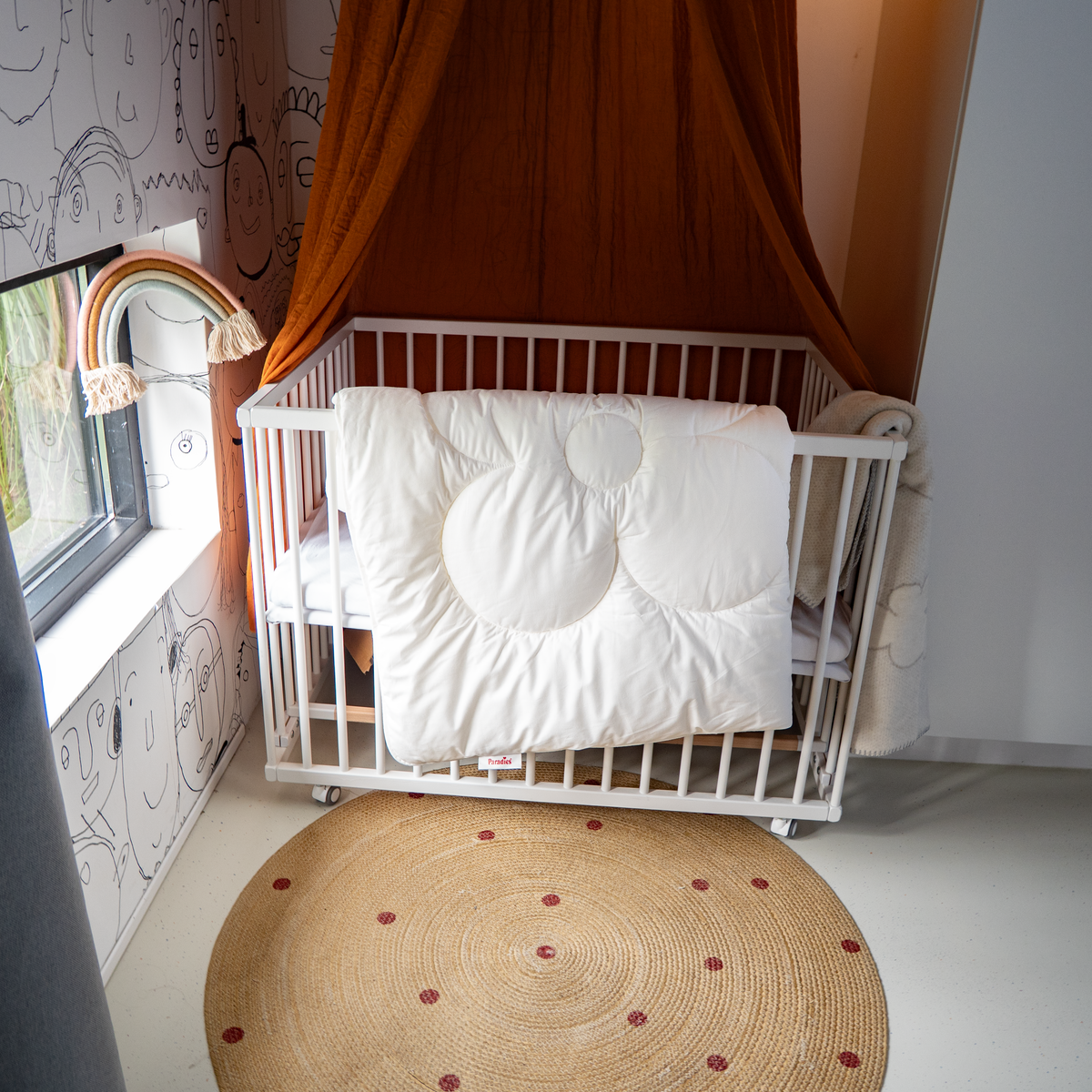 Kinderbett mit weißer Steppdecke mit großen runden Steppungen in einem Zimmer mit gemusterter Tapete, rundem Teppich und Regenbogen-Wandbehang