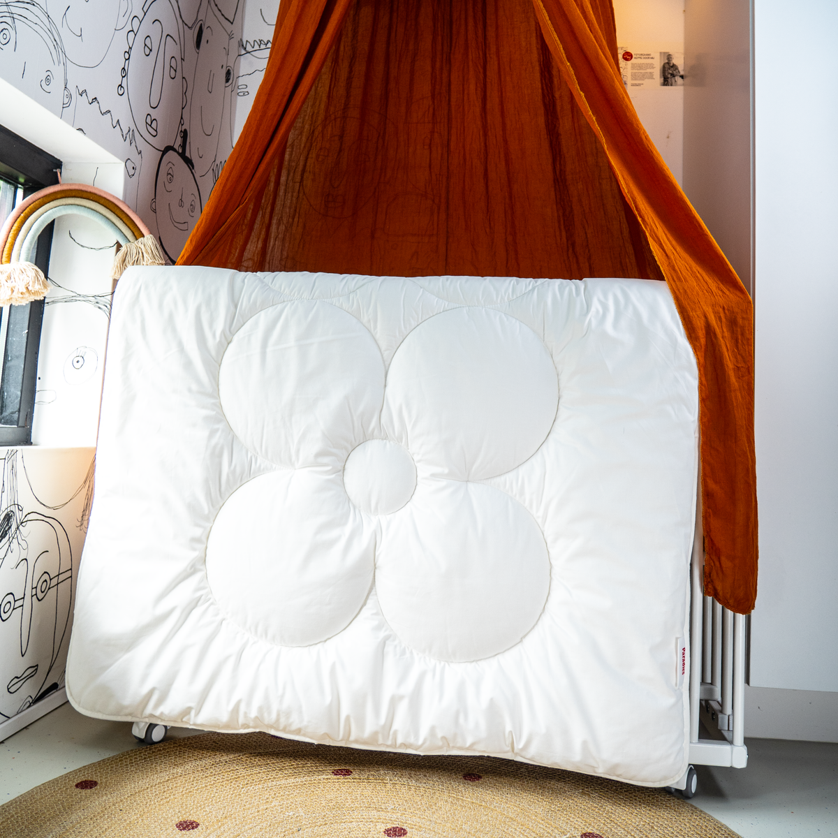 Weiße Kinderbettdecke mit großer, gesteppten Blütenform lehnt an einem orangefarbenen Baldachin in einem Kinderzimmer