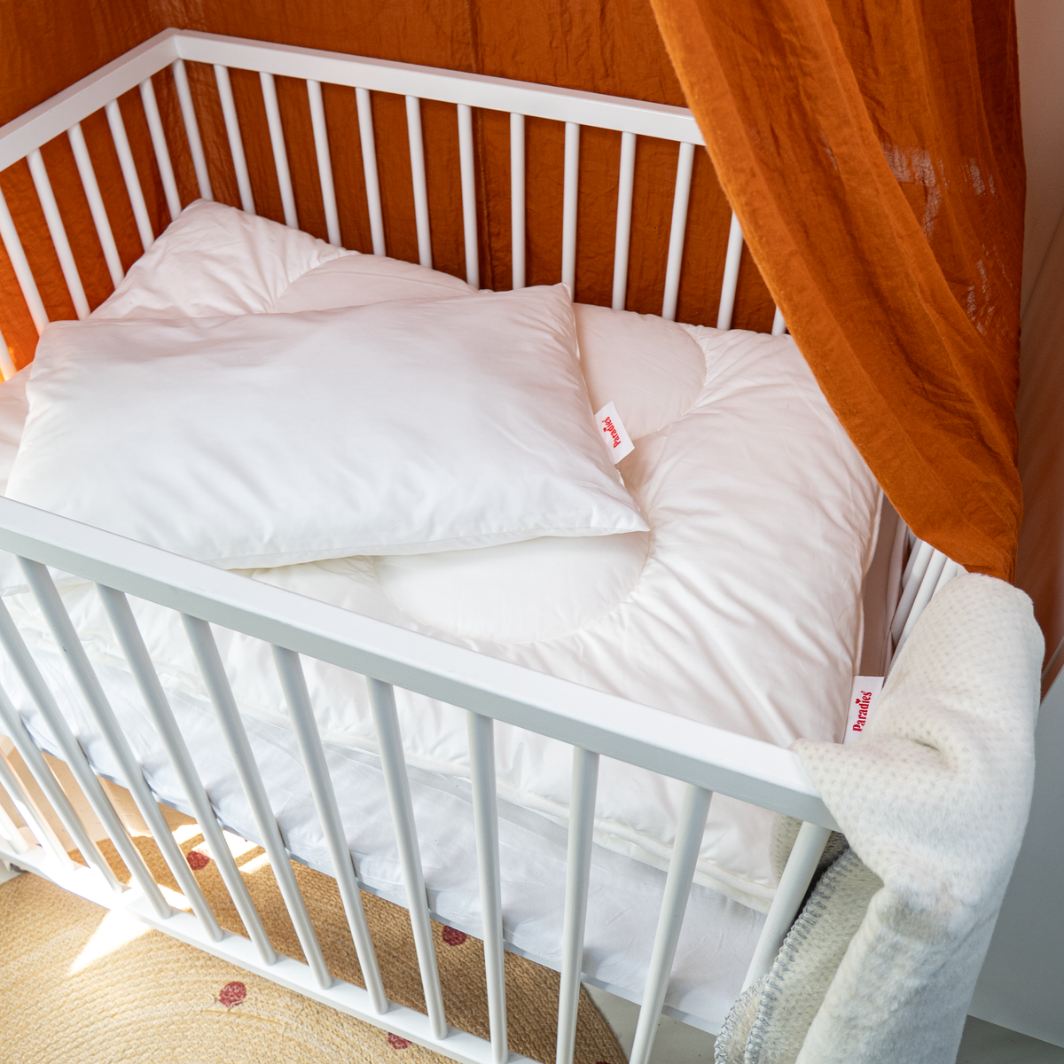 Kinderbett mit weißer Decke und Kissen, umgeben von orangefarbenem Vorhang 