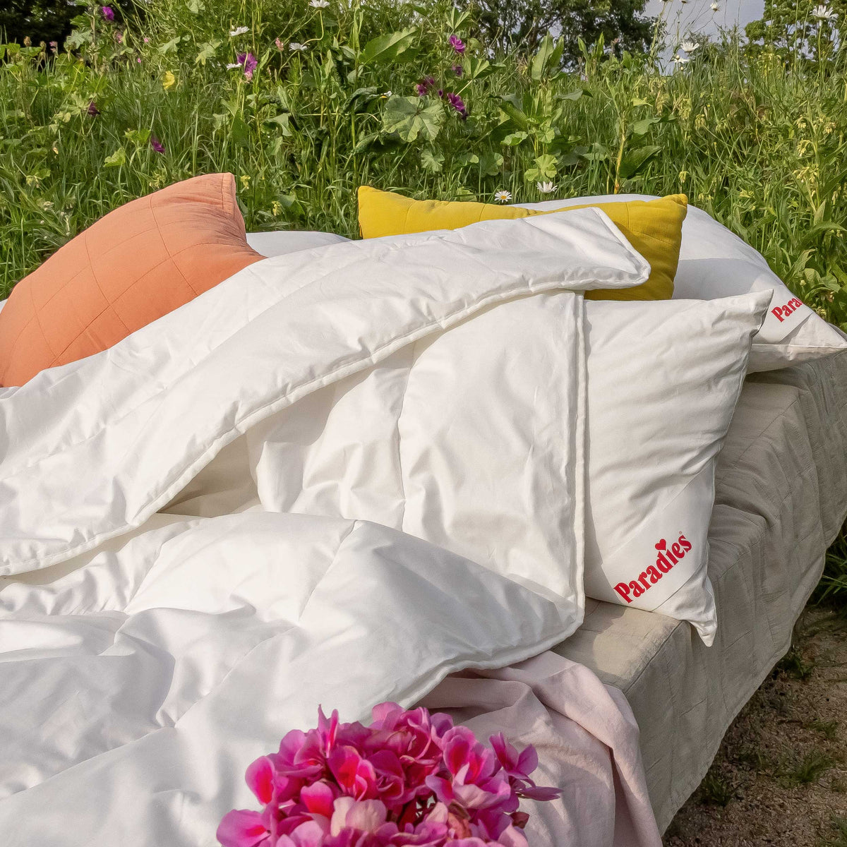 Bett mit weißen Kissen und Decke mit rotem Paradies-Logo vor einer Wiese mit grünen Pflanzen und Blumen