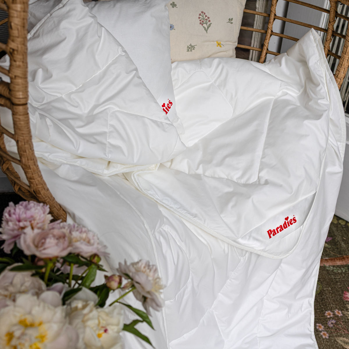 Weiße Prima Sommerbettdecke liegt in einem Korbsessel, daneben Blumen und ein Kissen mit floralem Muster