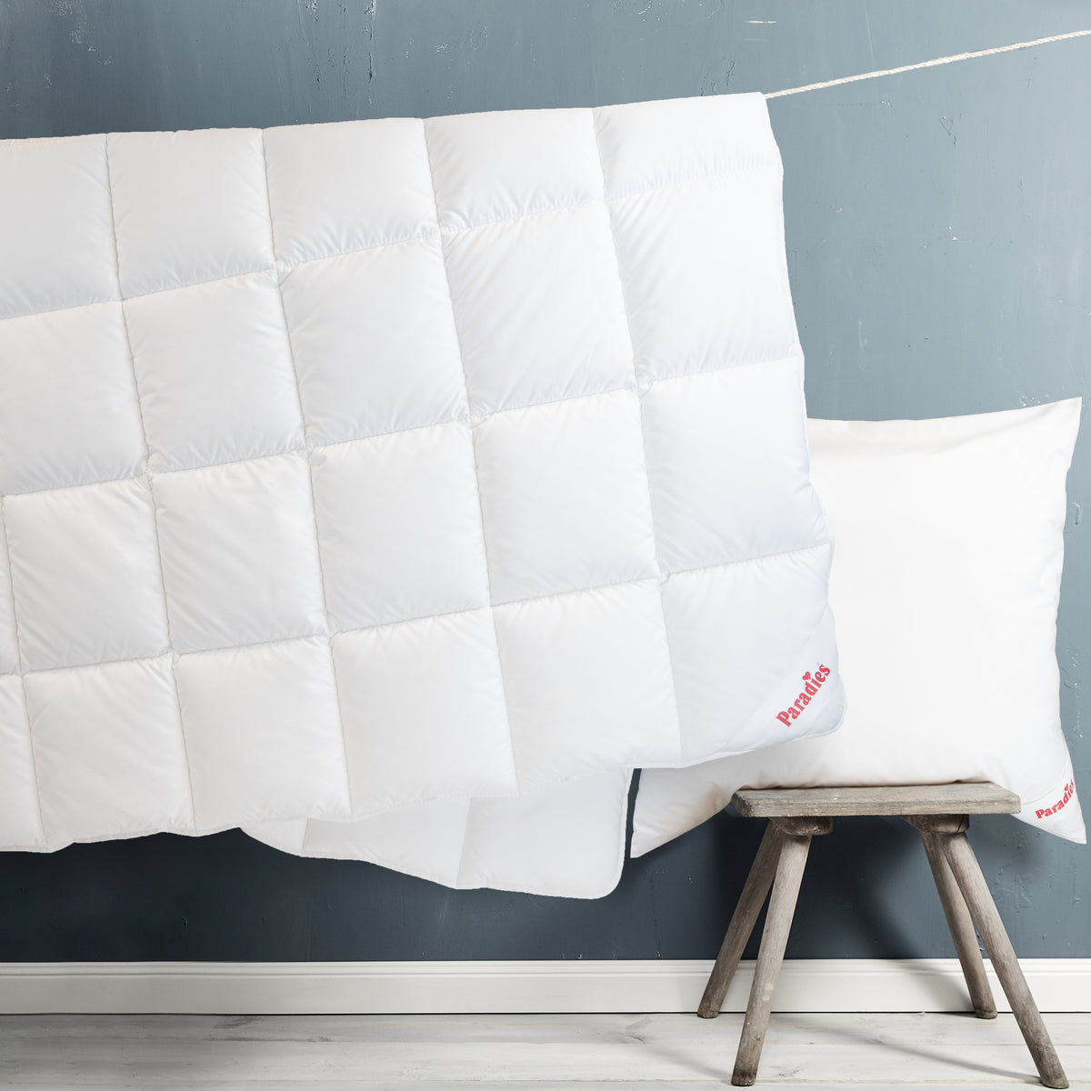 Großes weißes Steppbett hängt an einer Leine vor einer blauen Wand neben einem kleinen Holzschemel mit einem weißen Kissen
