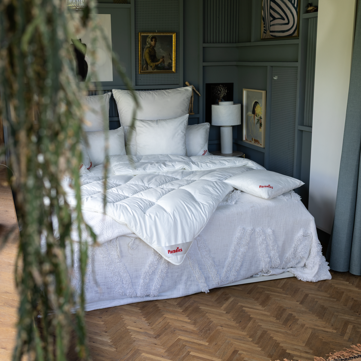 Weiße Paradies Runa Ganzjahresbettdecke mit Bodyform-Steppung auf Bett mit weißen Kissen in Schlafzimmer. Im Vordergrund hängt eine Pflanze ins Bild
