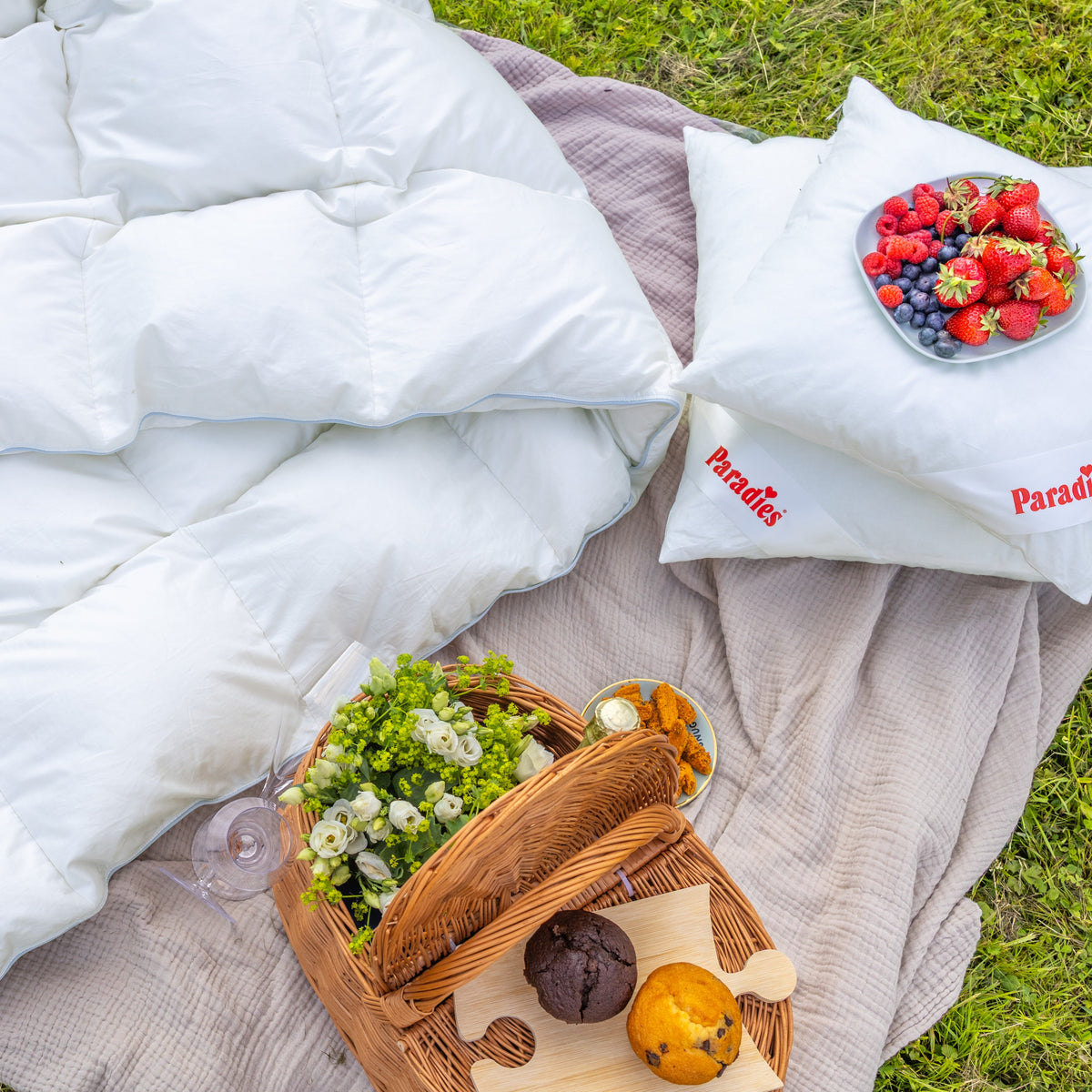 Picknickdecke auf Gras mit weißer Daunendecke, zwei Kissen mit rotem Schriftzug "Paradies", Korb mit Blumen und Gebäck sowie Teller mit Beeren