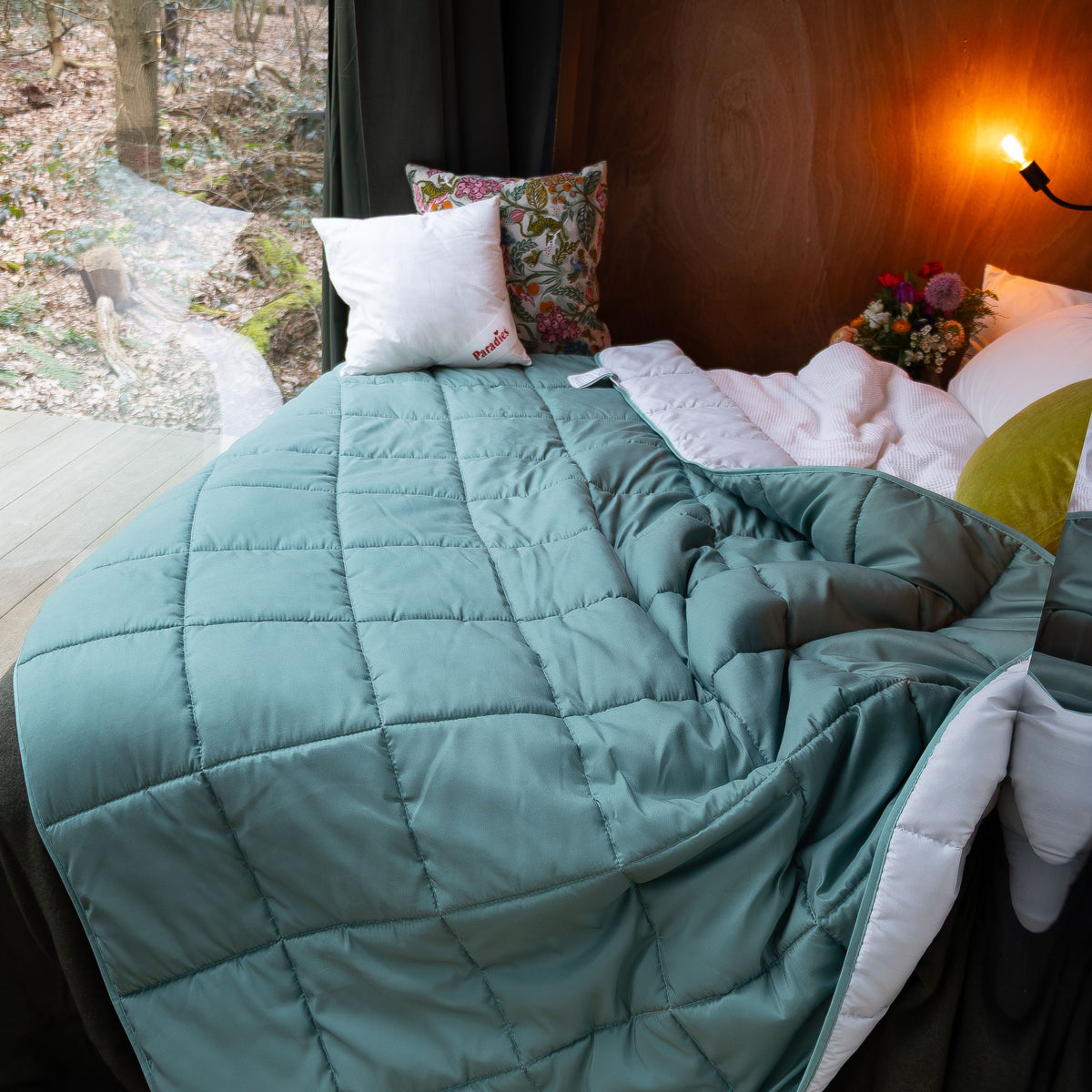 Bett mit hellgrüner Steppdecke Sorelia, mehreren Kissen und Blick auf einen Wald durch ein Fenster