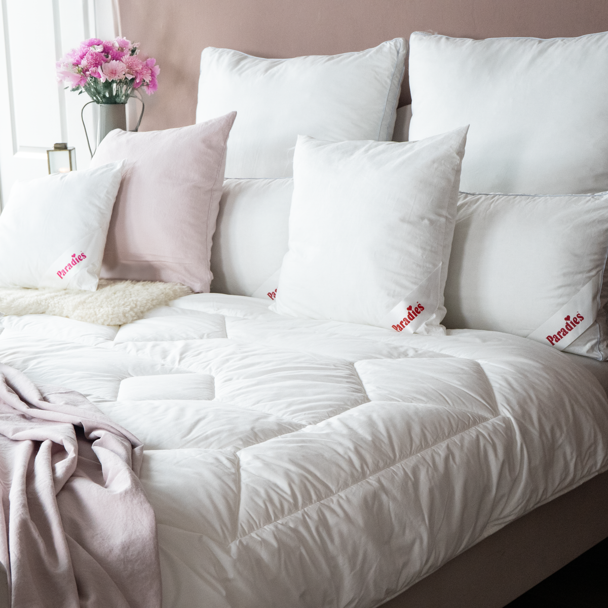 Bett mit weißer Steppdecke, mehreren weißen und einem rosafarbenen Kissen mit roter Aufschrift sowie einem Blumenstrauß in Vase auf Nachttisch
