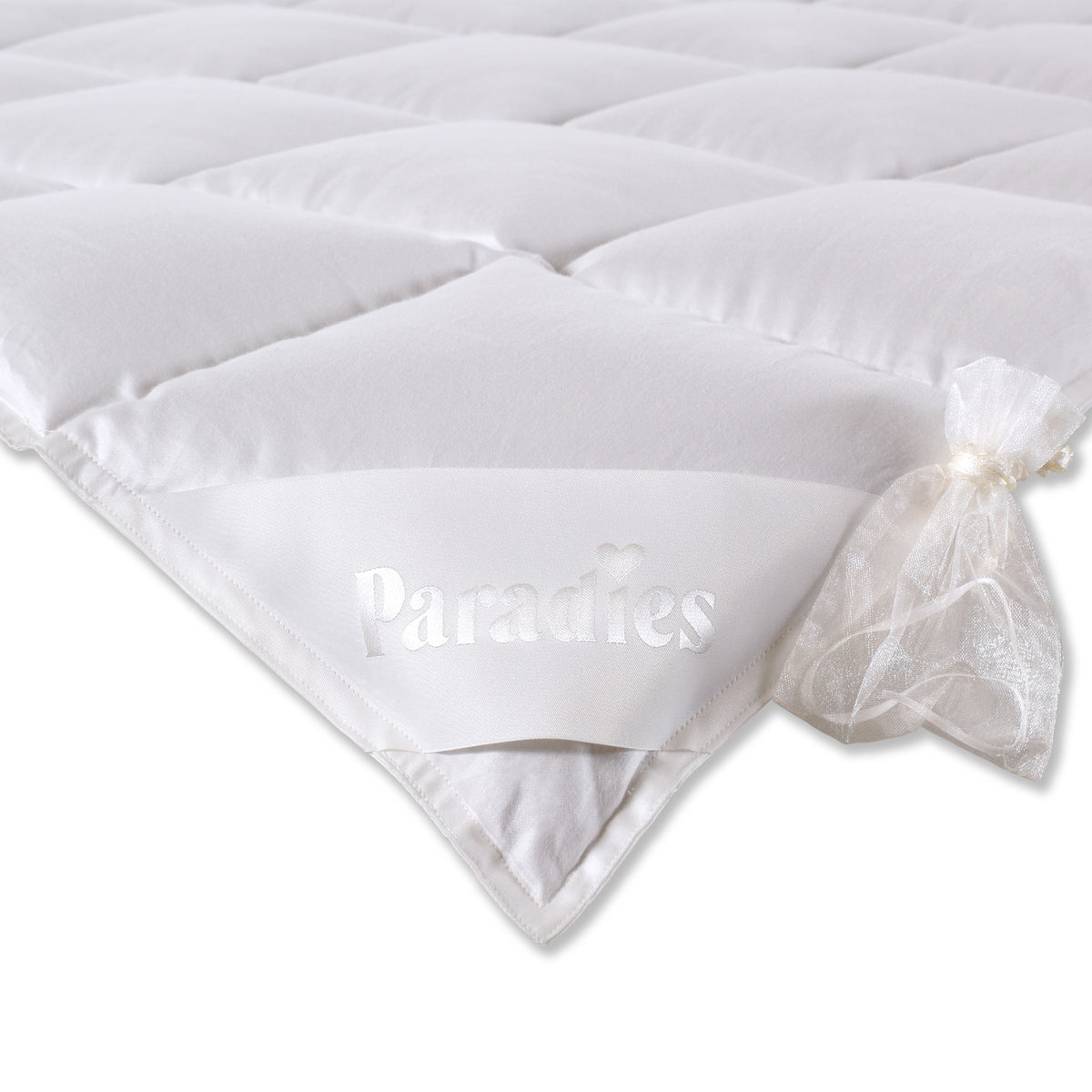 Nahaufnahme einer weißen Steppdecke mit dem Schriftzug 'Paradies' und einem kleinen weißen Organza-Beutel an der Ecke